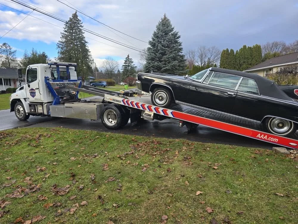 A tow truck carrying a black vintage car on a suburban street with houses, trees, and a cloudy sky in the background.