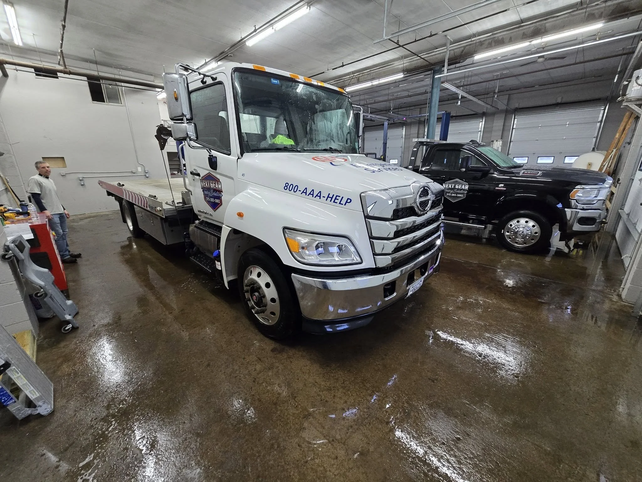 Inside a garage with a flatbed tow truck, a black pickup truck, and a person standing near the wall.