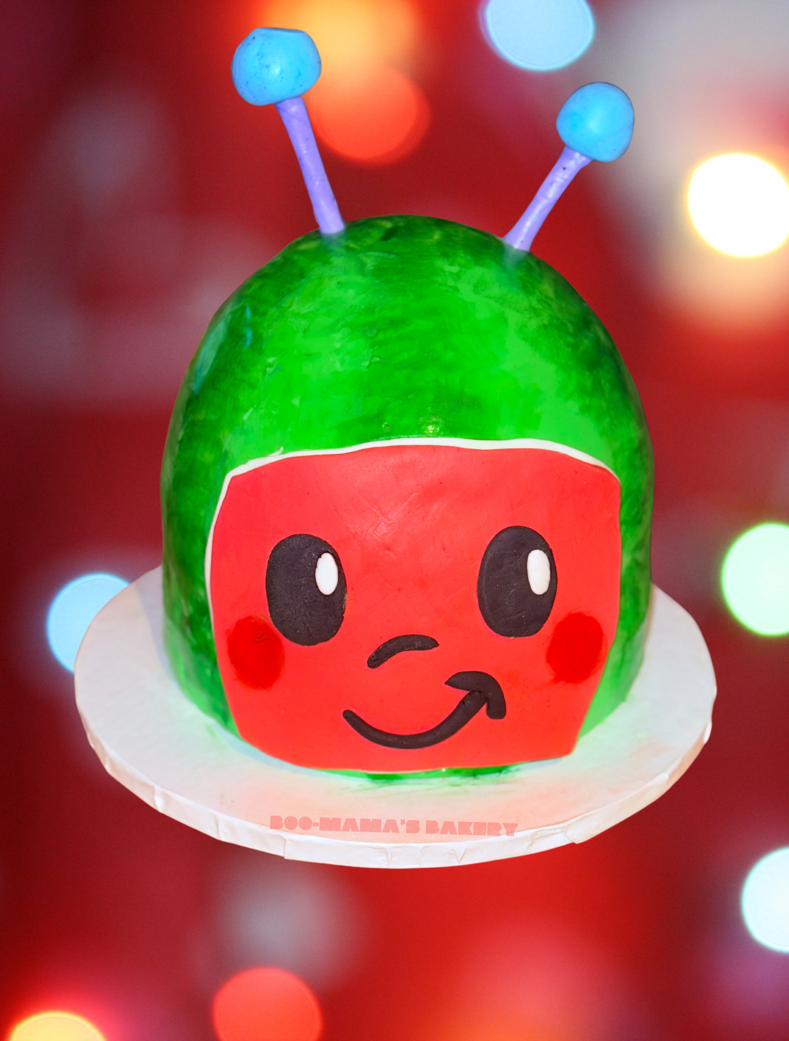 A fruit-themed cake decorated to resemble a cartoon ladybug with a smiling face, antennae, and a red face with black eyes and a mouth, on a white cake base with the text 'BOO-MAMA'S BAKERY'.