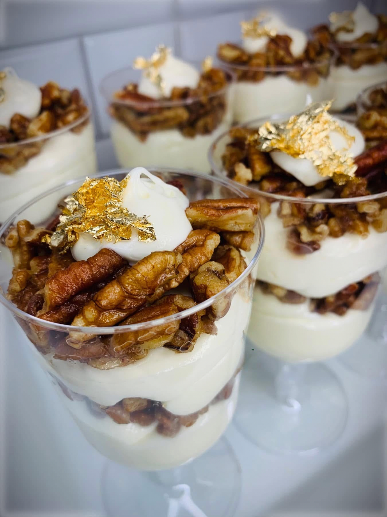 Dessert parfait in clear glasses layered with whipped cream, chopped pecans, and topped with edible gold flakes.