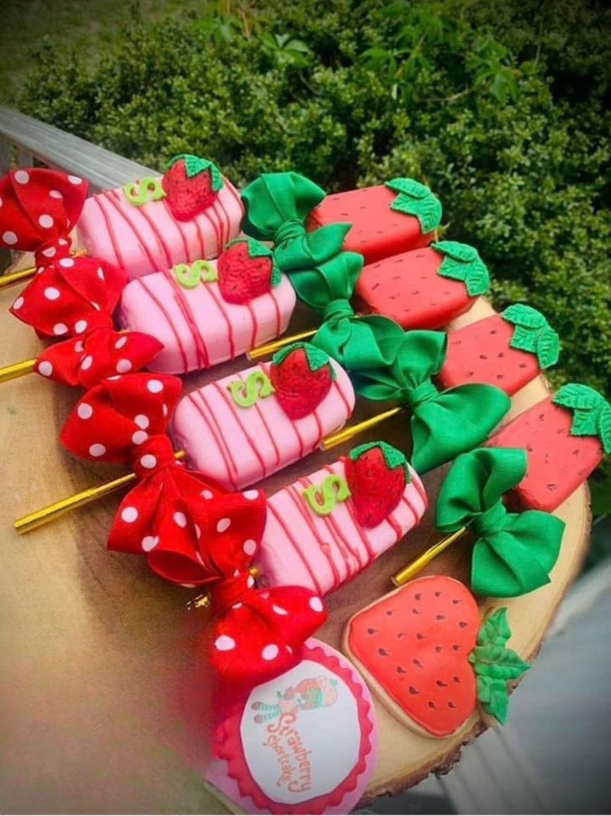 Decorative strawberry-shaped cookies with pink and red icing, green ribbon bows, and small strawberry and dollar sign decorations, arranged on a wooden surface against a green hedge background.