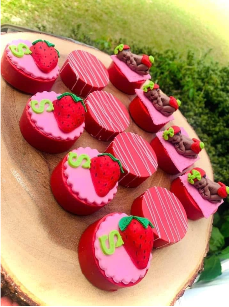 A wooden serving platter with strawberry-themed cupcakes and figurines, accompanied by decorative cake boxes, set outdoors with greenery in the background.
