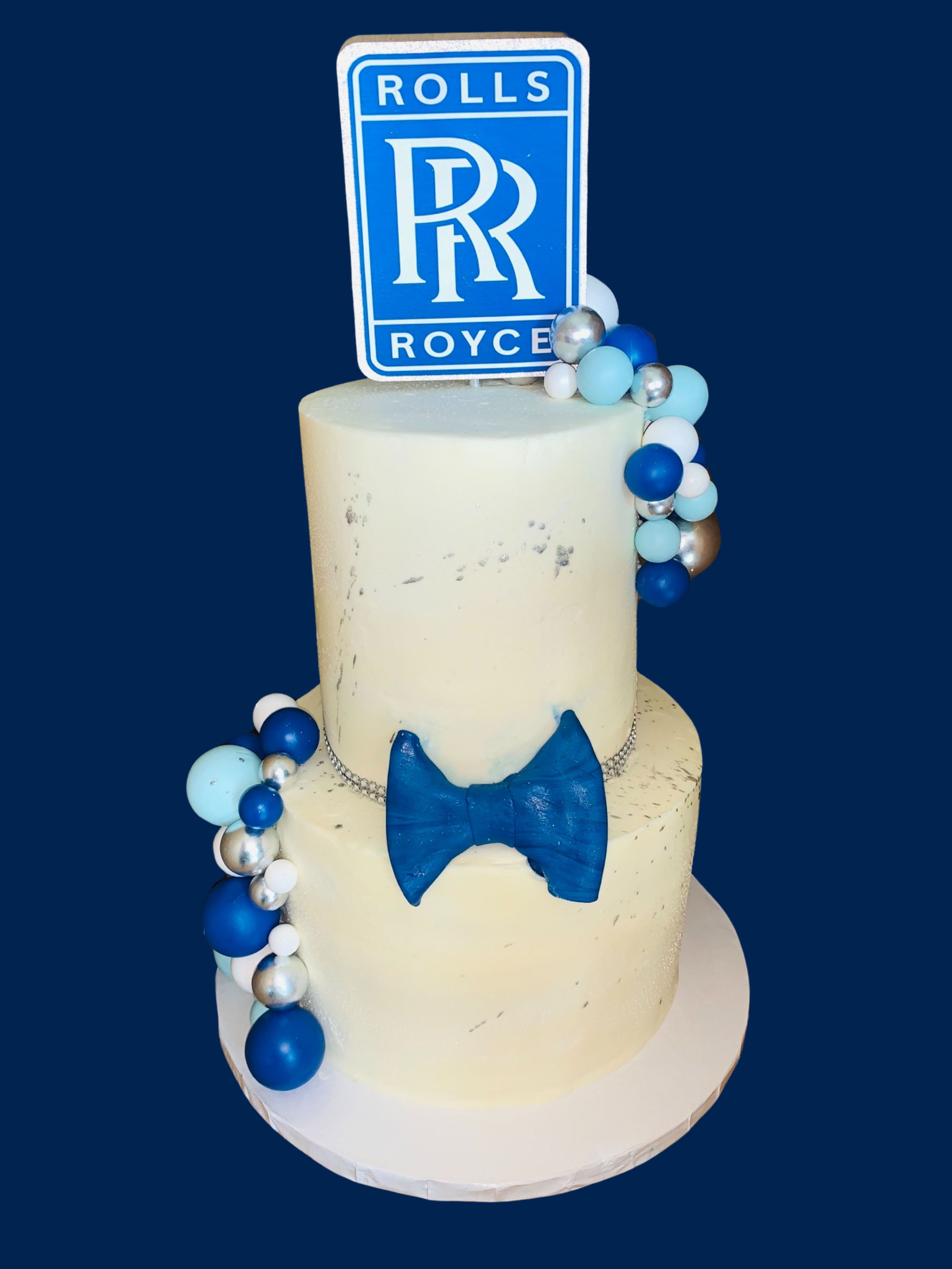 Two-tiered cake decorated in blue and white with a Rolls Royce logo topper and a blue bowtie on the front.
