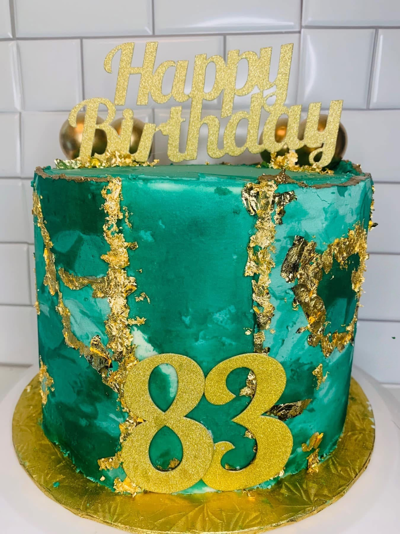 A turquoise birthday cake decorated with gold foil accents, a large gold glitter number "83", and a gold "Happy Birthday" topper, placed on a gold cake board.