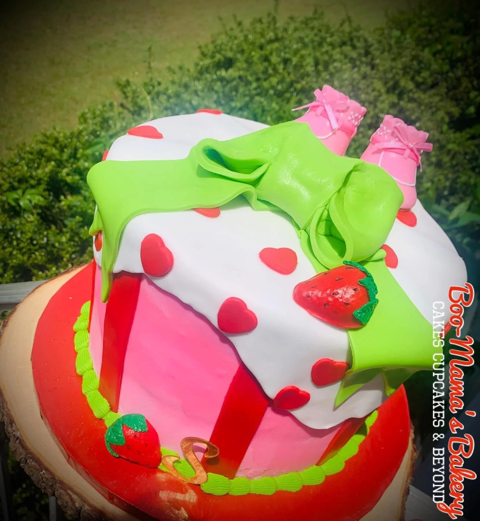 A cake designed to look like a strawberry-themed gift box, decorated with pink and green fondant, strawberry-shaped fondant accents, and pink fondant baby shoes on top, set against a outdoor background.