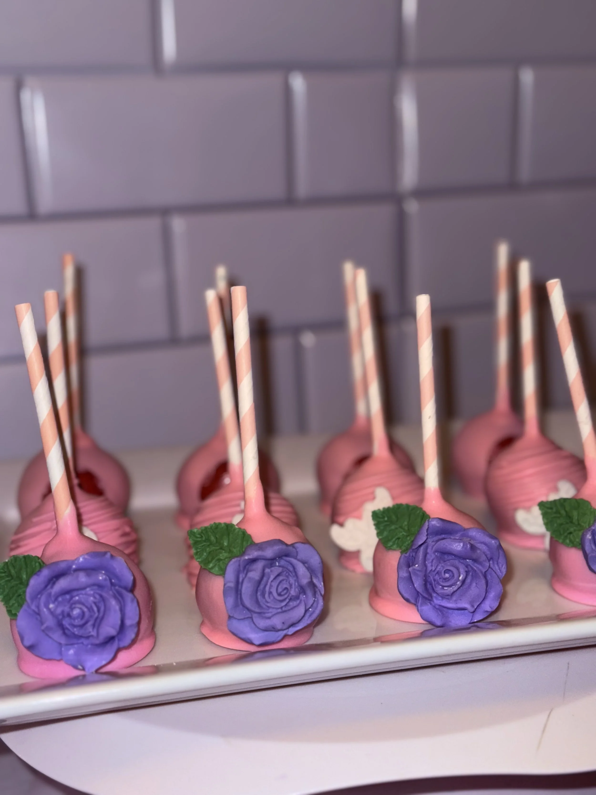 Pink cake pops decorated with purple roses and green leaves, each with a striped pink and white straw, lined up on a white tray against a tiled gray wall.