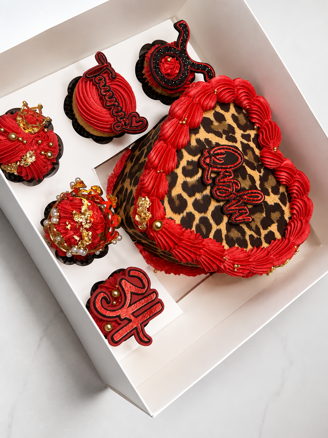A white gift box contains a leopard print cake with red frosting, decorated with gold accents and a red "Happy" topper. Surrounding the cake are six decorated cupcakes with red frosting, gold embellishments, and themed toppers, some reading "Thankful