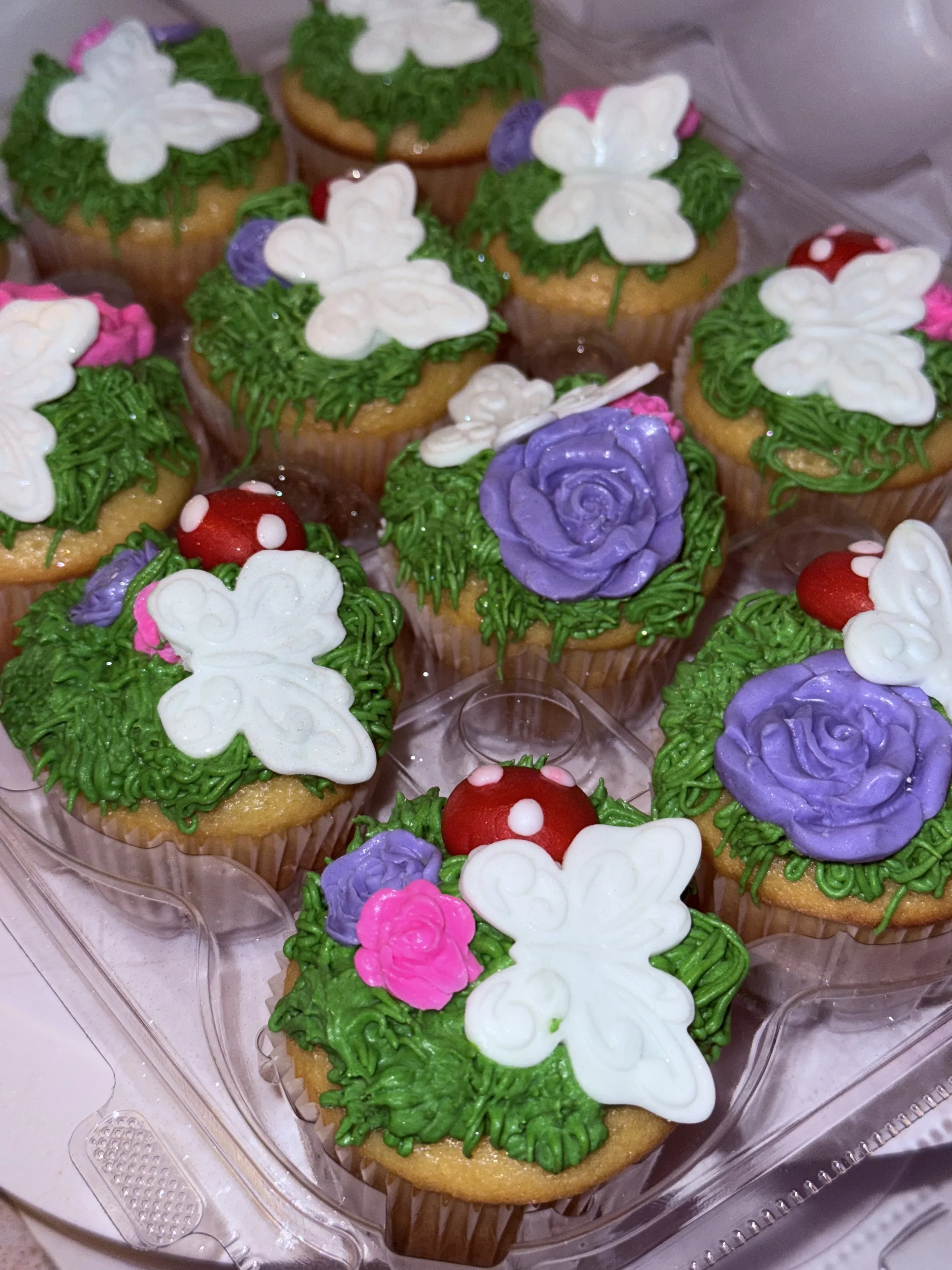 Decorated cupcakes with green frosting, white butterfly shapes, purple roses, red and white mushroom-shaped candies, and pink floral details in a plastic container.