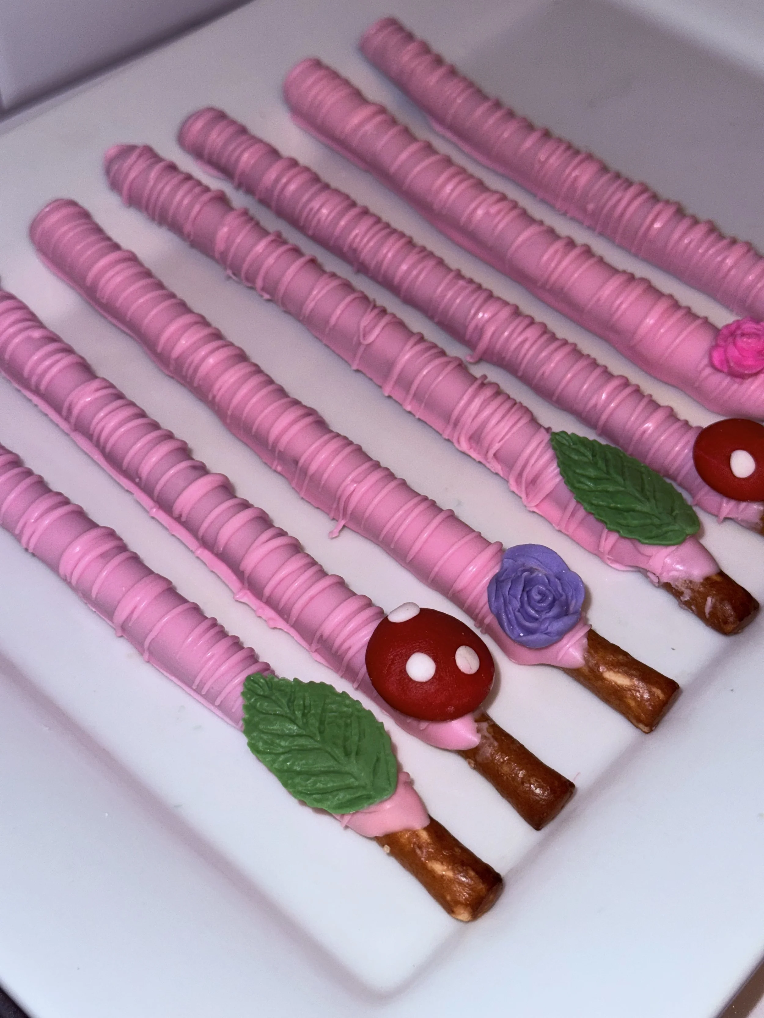 Pink decorated pretzel sticks with colorful toppings and leaves arranged on a white tray.