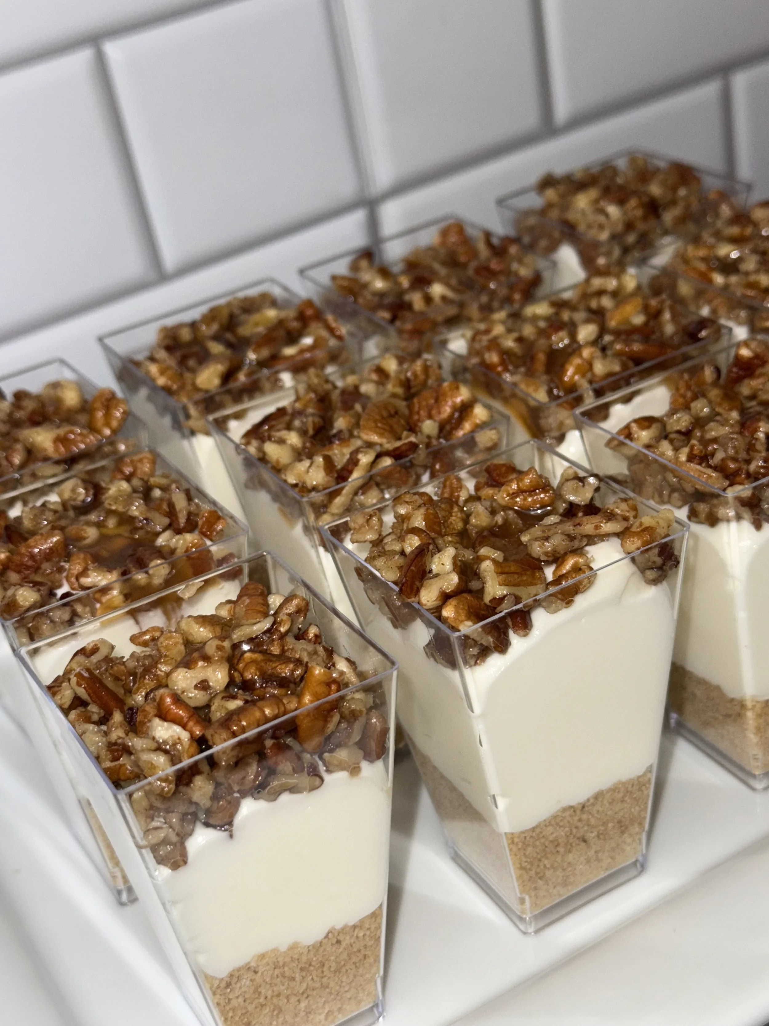 Individual layered desserts in clear cups, with a graham cracker crust, a cream cheese filling, and topped with chopped pecans.