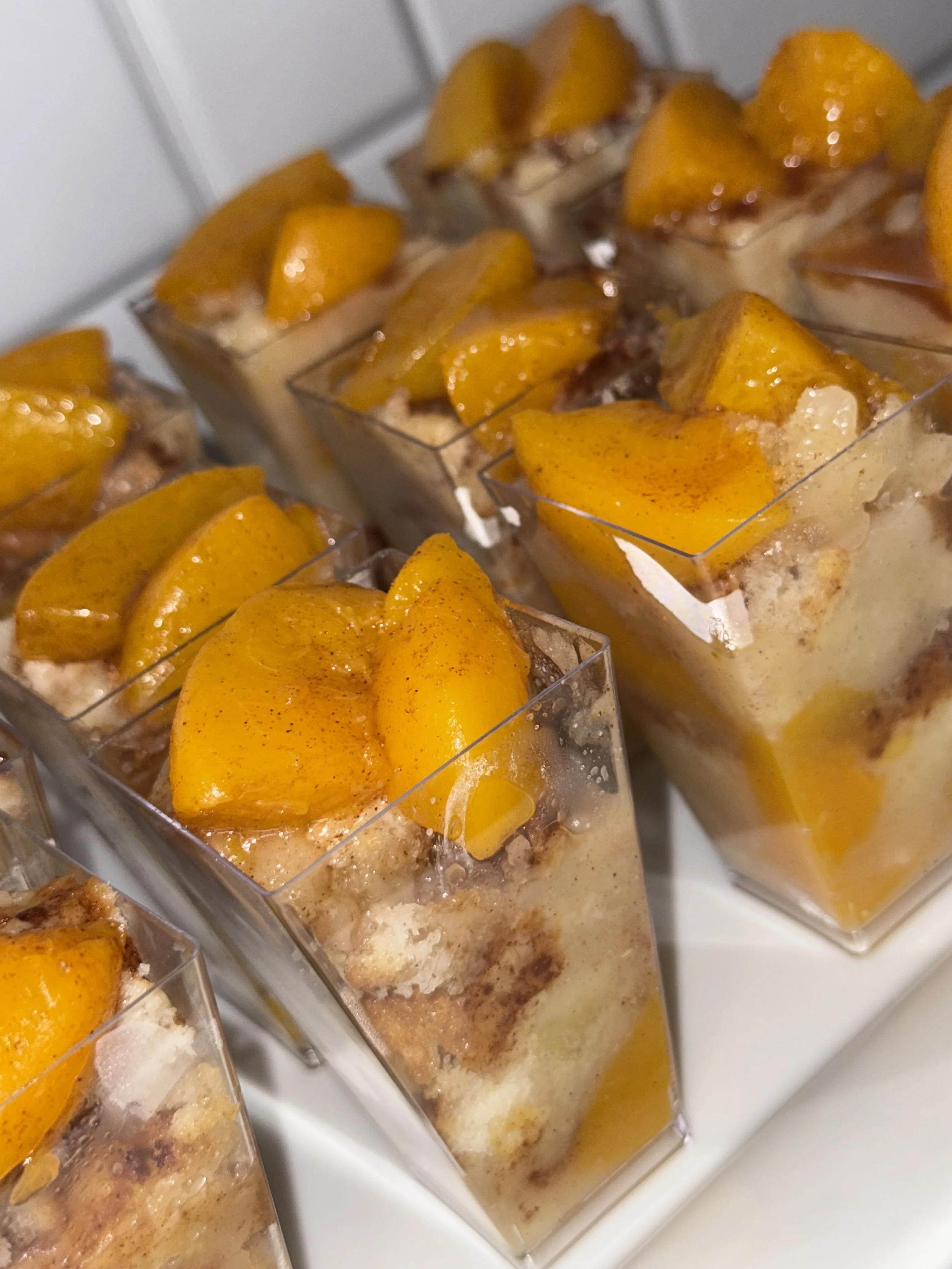 Close-up of individual dessert parfaits with layers of cream, crumbled biscuits, and topped with fresh peach slices in clear square cups.
