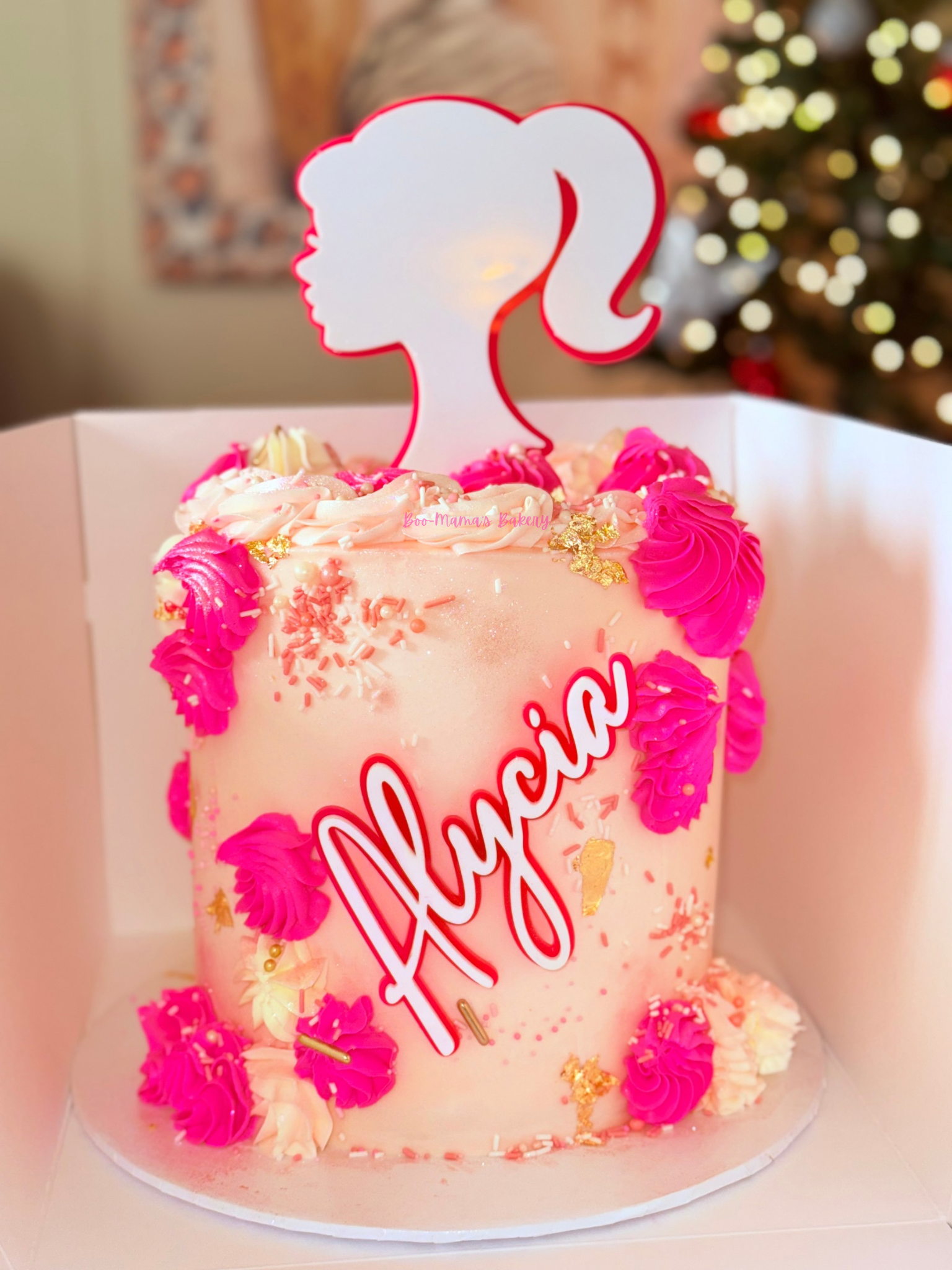 Pink birthday cake decorated with pink and white frosting, gold leaf, sprinkles, and a silhouette of a girl's face with a ponytail on top. The cake has a sign that says "Alycia" and a gold number "7" for the age, set against a background with a Chris