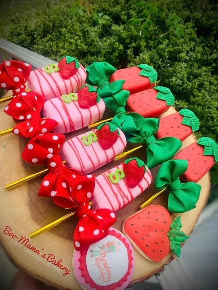 Assorted strawberry-themed cake pops with bows and strawberry decorations on a wooden platter, labeled "Boo Mama's Bakery" and a tag that says "Sunshine & Smiles".
