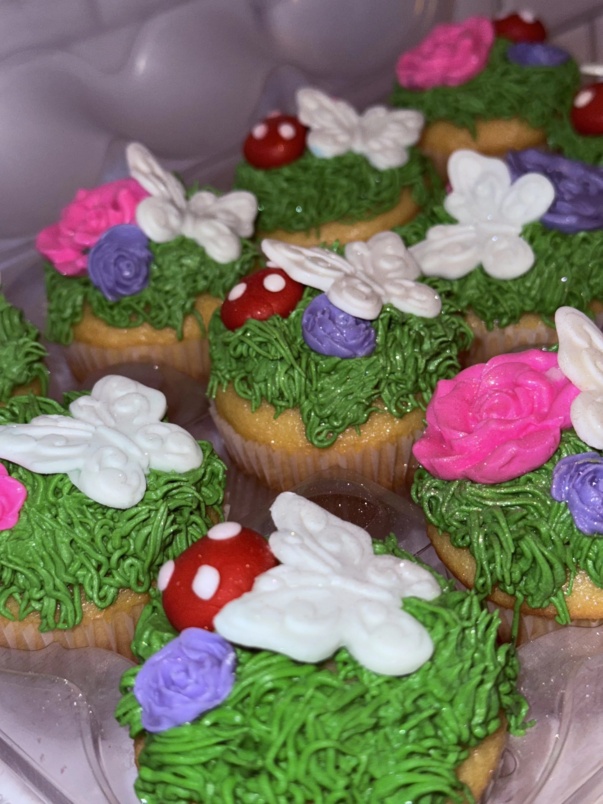 Decorated cupcakes with green frosting, white, pink, and purple flowers, white butterflies, and red mushroom toppers.