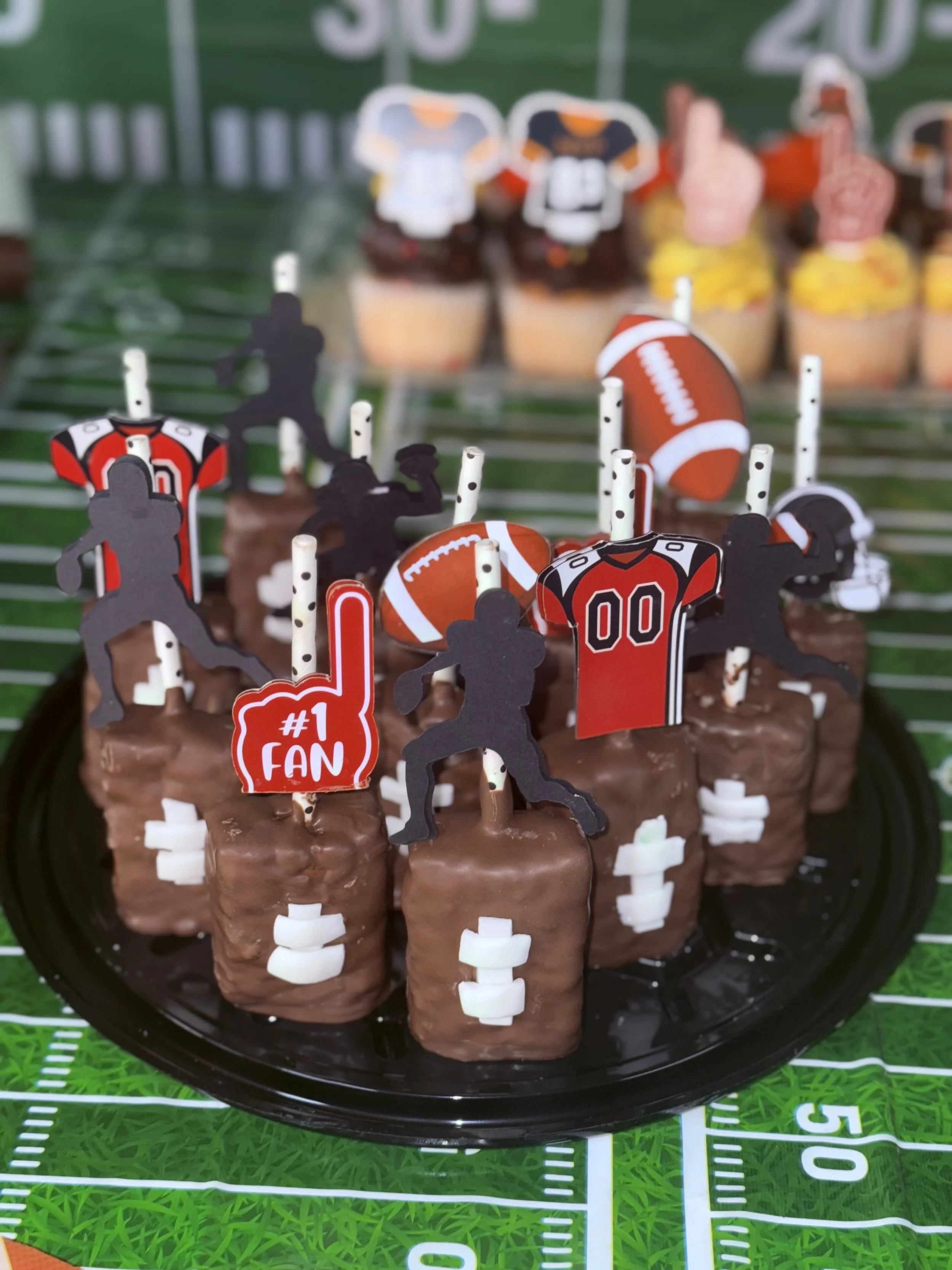 Football-themed chocolate cupcakes with football and player toppers, including a red foam finger that says '#1 FAN' and mini foam footballs, arranged on a black platter in front of a football field-themed tablecloth.