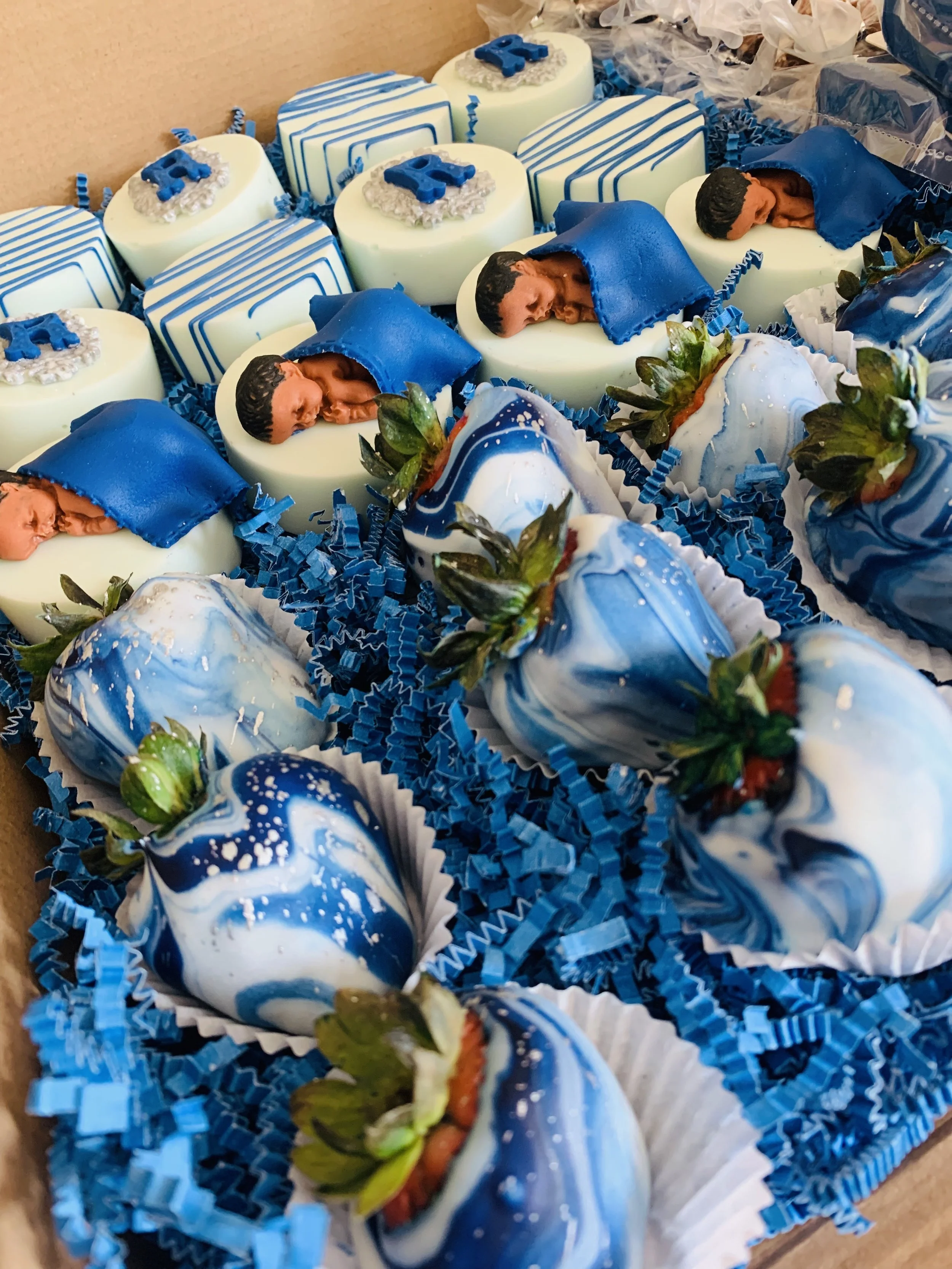 Assorted decorated chocolates, including some shaped like strawberries and others with cake topper designs, arranged on blue shredded paper in a box.
