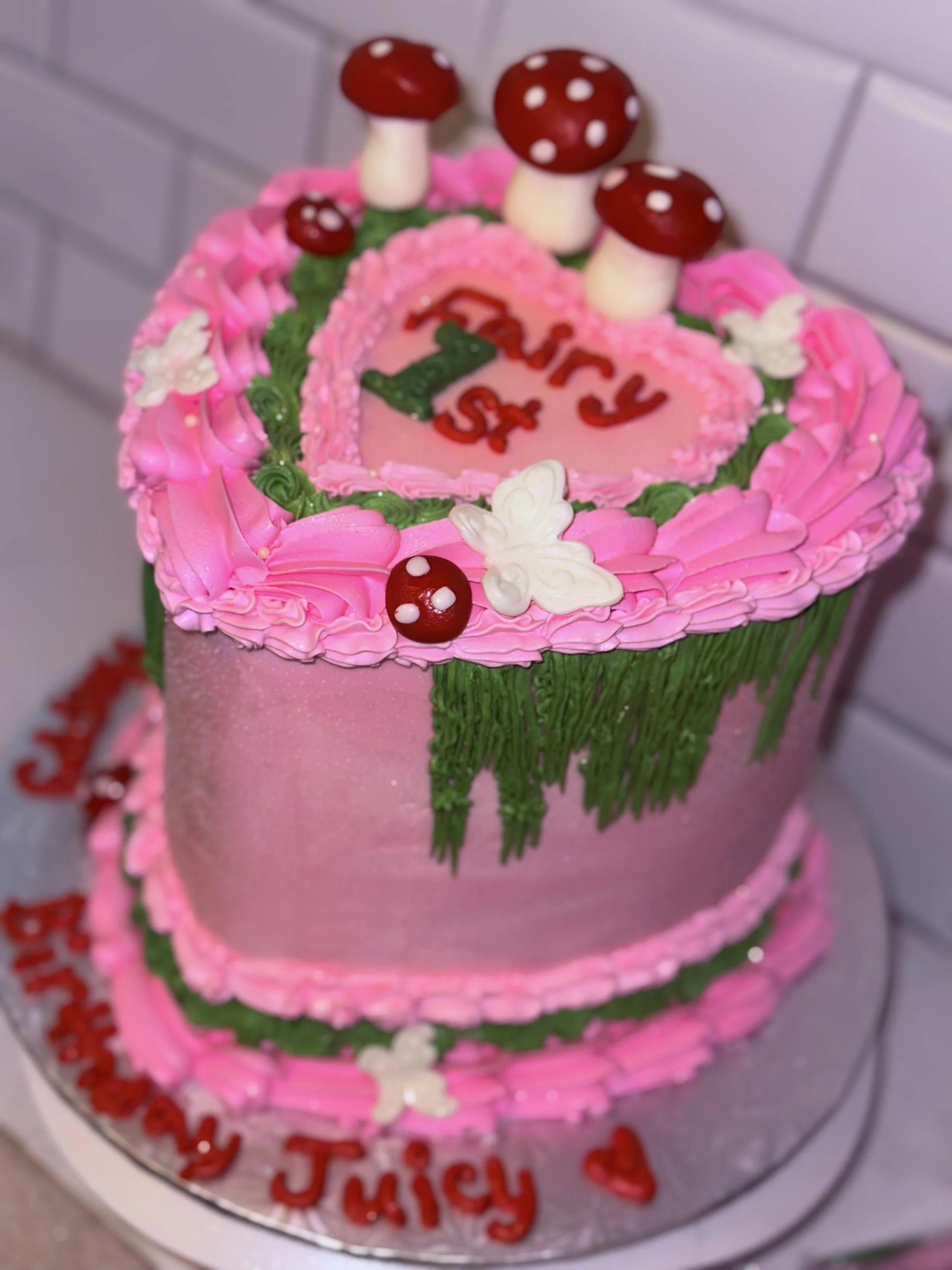 A two-tier pink birthday cake decorated with pink, green, and white frosting, topped with red and white mushrooms, white butterfly decorations, and the message "Happy 5th JiuJY" written in red on the cake board.