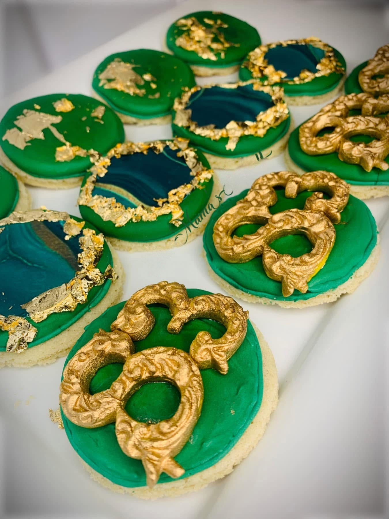 Decorated cookies with green, gold, and navy blue icing, some featuring gold leaf, arranged on a white surface.