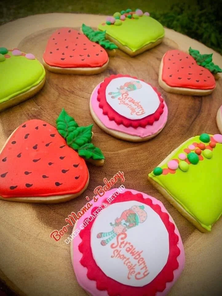 Decorated sugar cookies shaped and decorated like strawberries and strawberry plants on a round wooden surface, with colorful frosting and sprinkles.