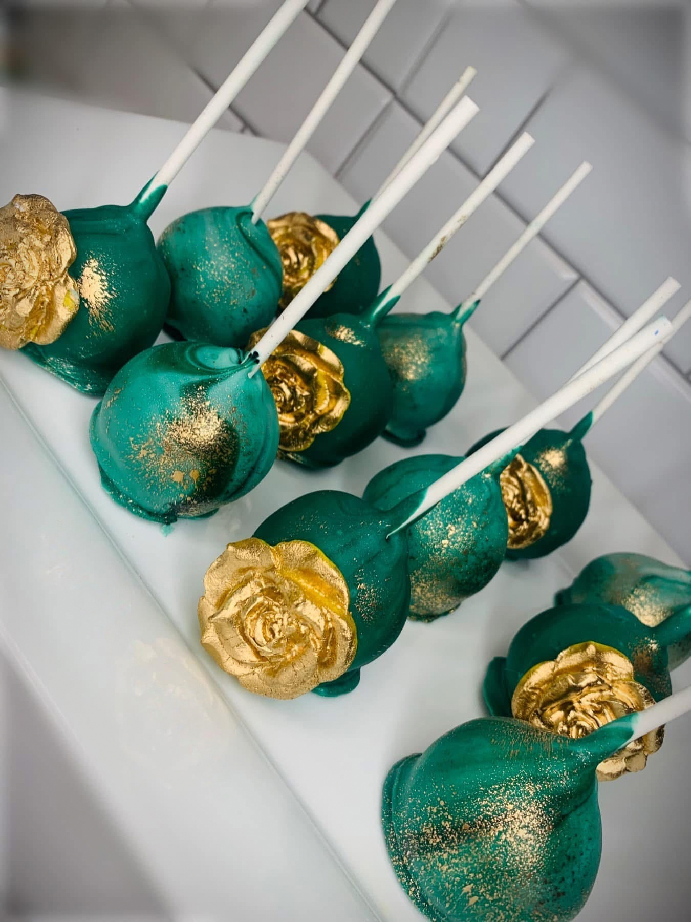 Cake pops decorated with teal and gold paint, resembling roses, on white sticks displayed on a white tray.
