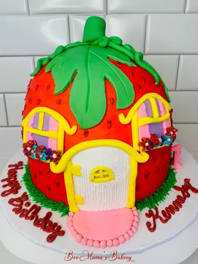 A large strawberry-shaped cake with green fondant leaves and stem, yellow and pink door, and arched windows decorated with colorful flowers, on a white cake board with a pink and red birthday message.