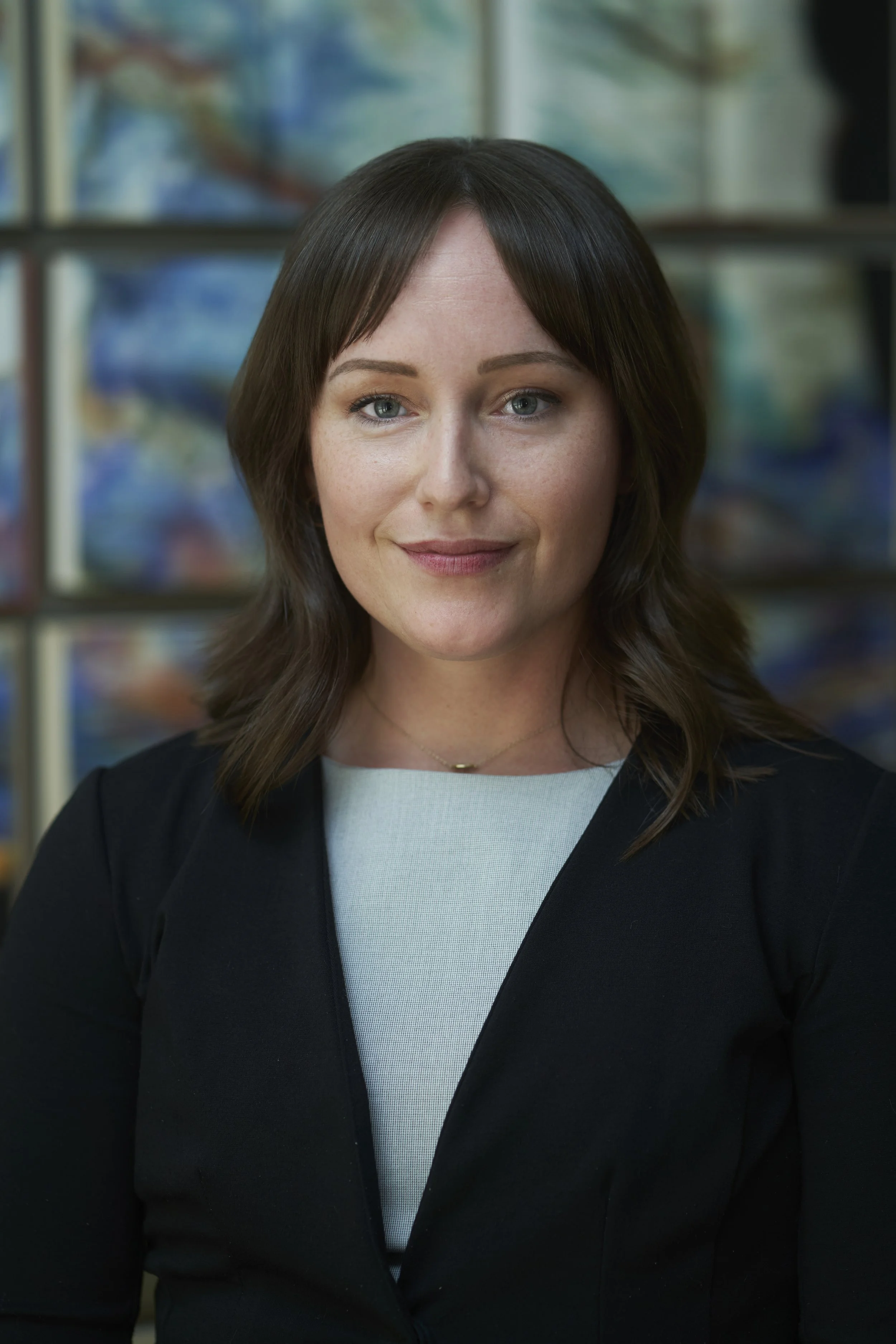 A woman with shoulder-length brown hair, wearing a black blazer over a white top, standing in front of a colorful abstract painting or mural.