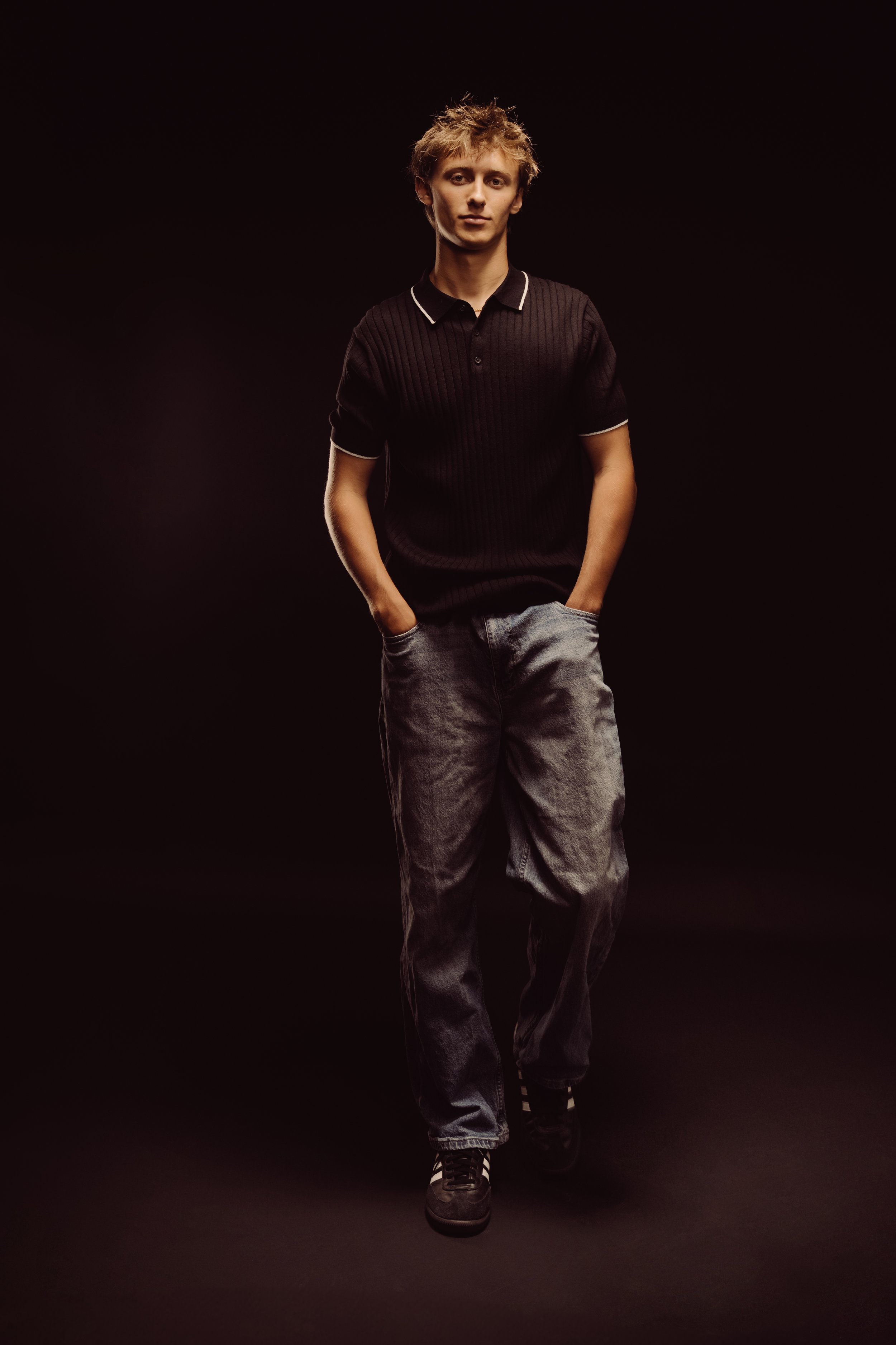 Young man walking forward with hands in pockets, dressed in a black polo shirt and loose gray jeans, against a dark background.