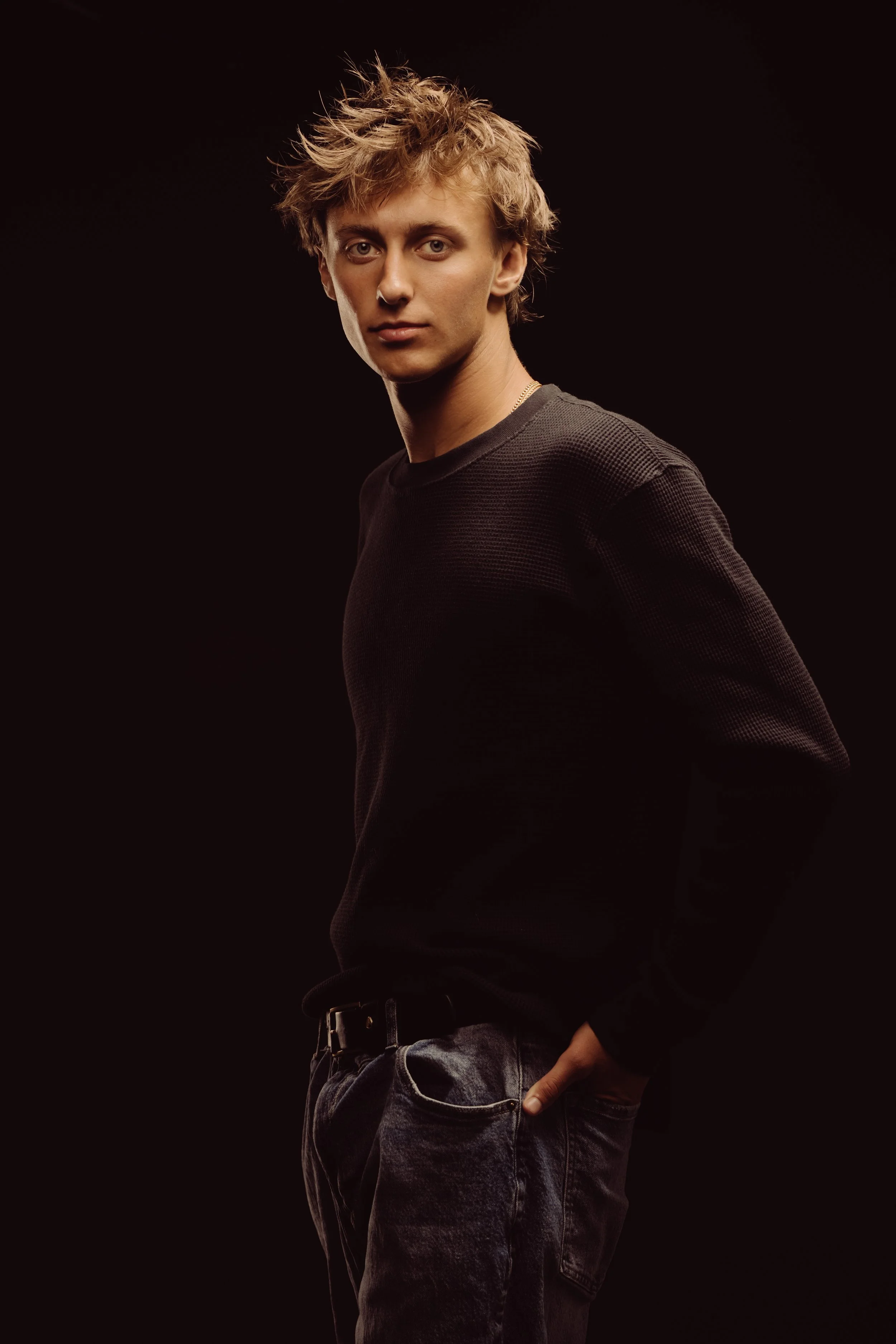 Young man with tousled hair wearing a black sweater and dark jeans, posing against a dark background.