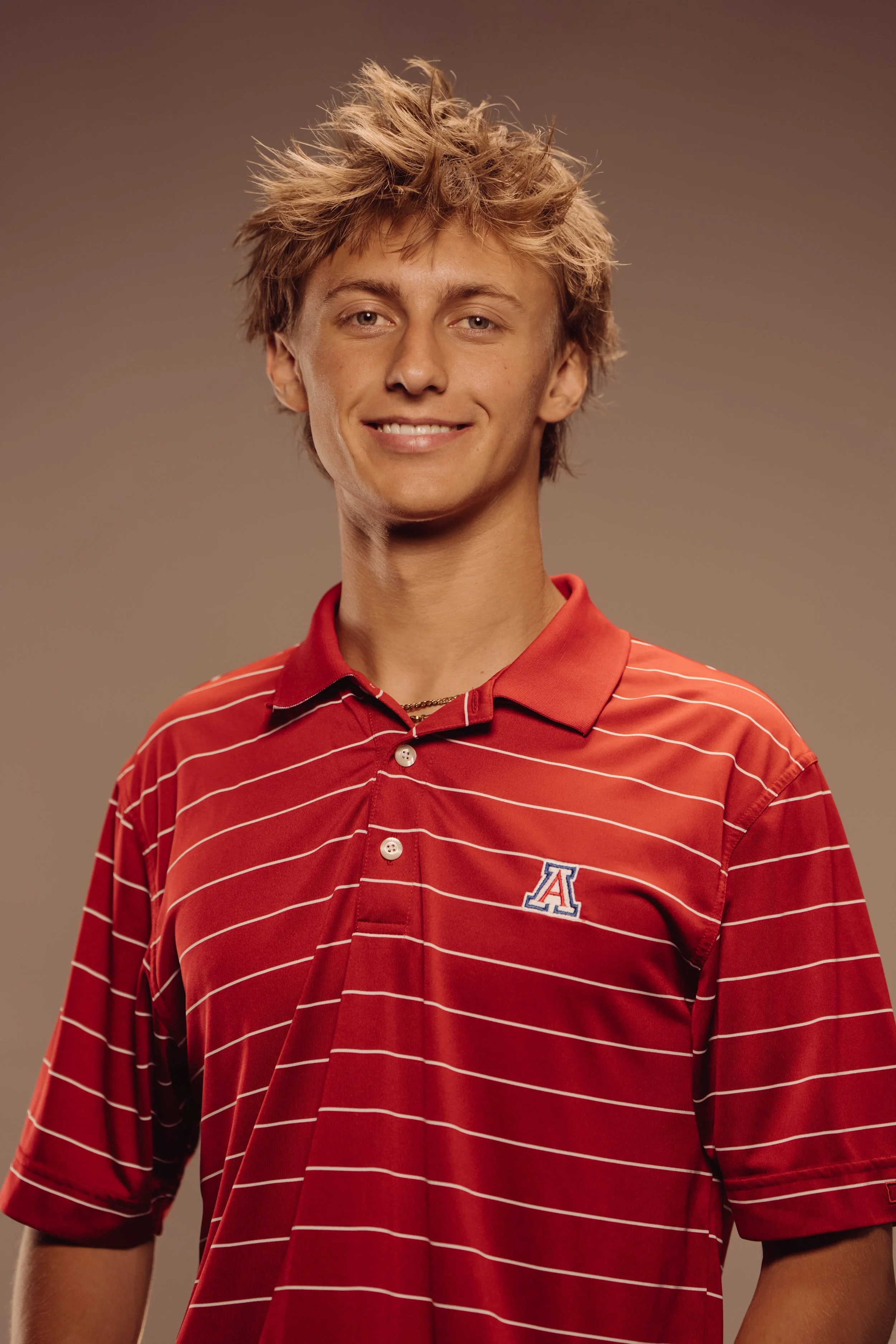 A young man with tousled blonde hair wearing a red striped polo shirt with an 'A' logo on the chest, smiling at the camera.