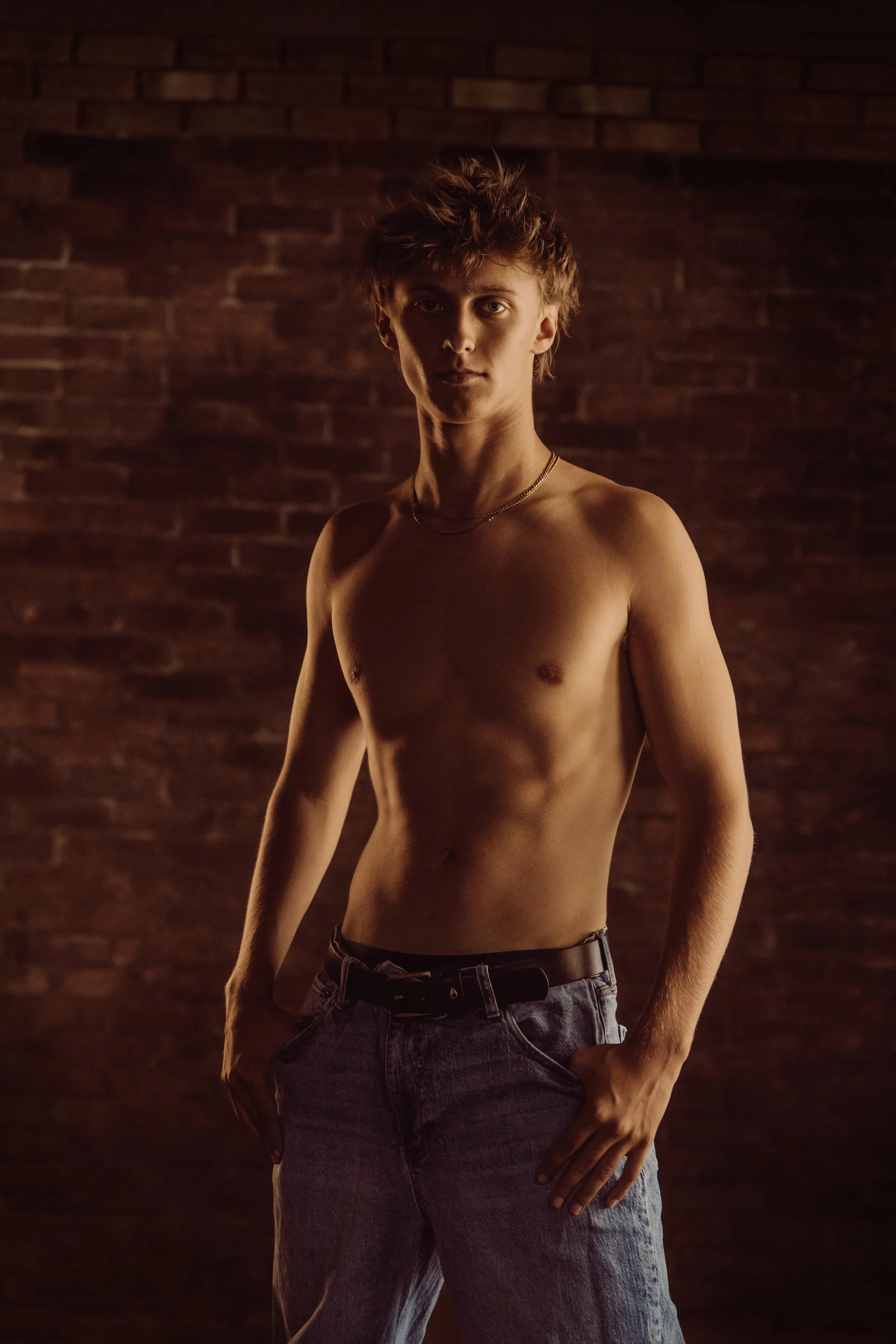 A shirtless young man with tousled hair and light skin standing against a dark brick wall.