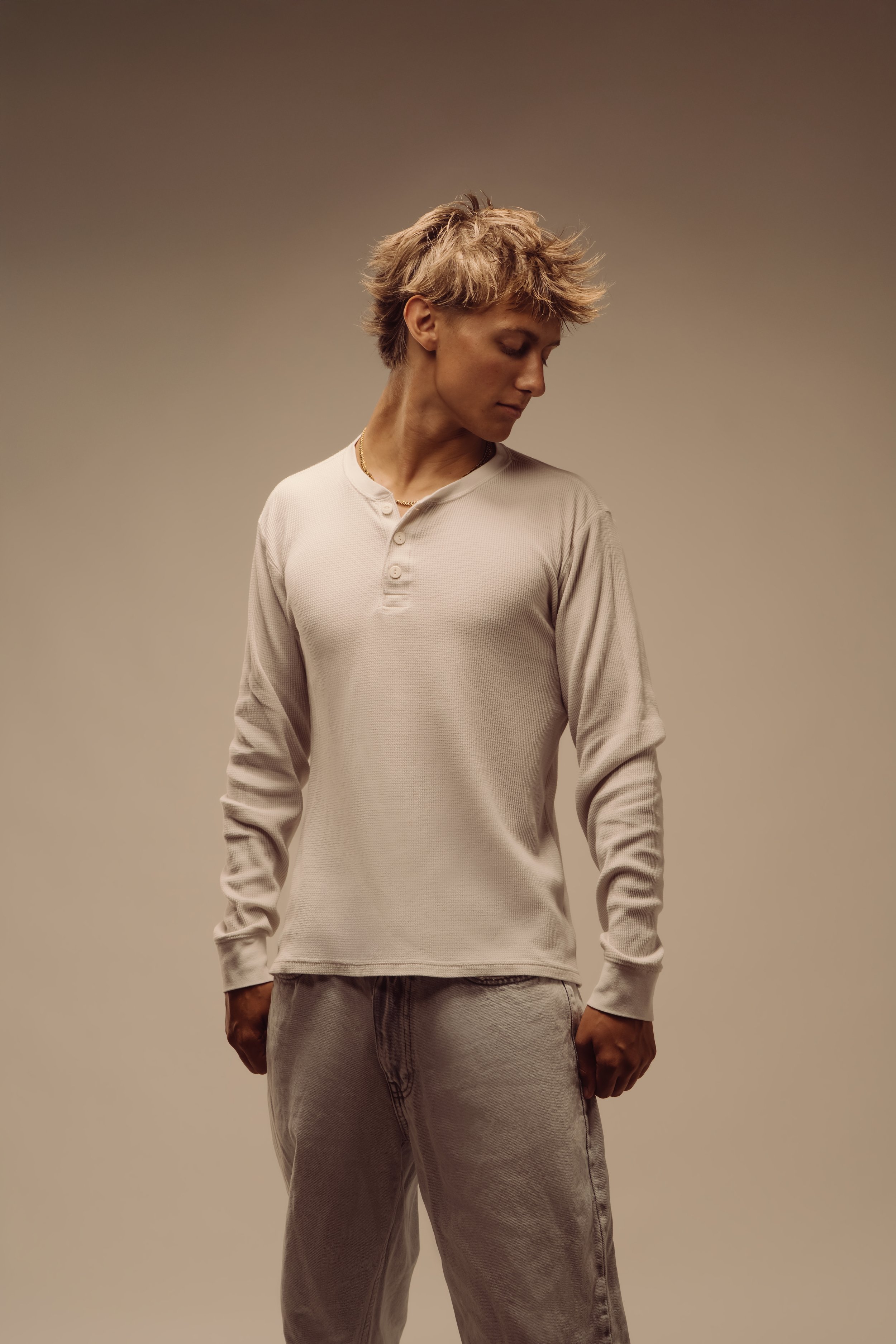 A young person with wavy blonde hair standing with a neutral expression, wearing a long-sleeved beige shirt and light-colored pants, in front of a plain background.