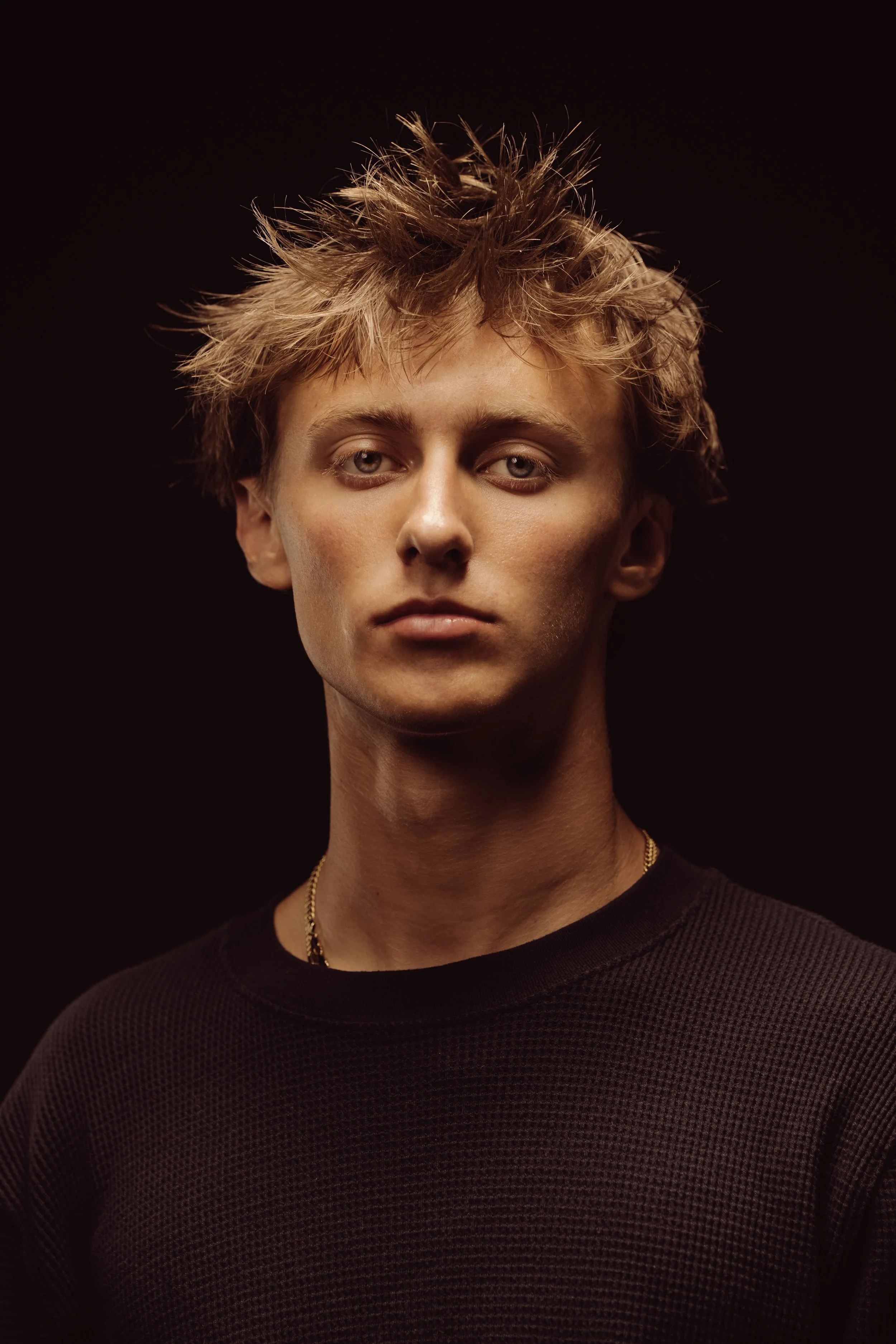 Portrait of a young man with messy blond hair, wearing a black textured shirt, against a dark background.