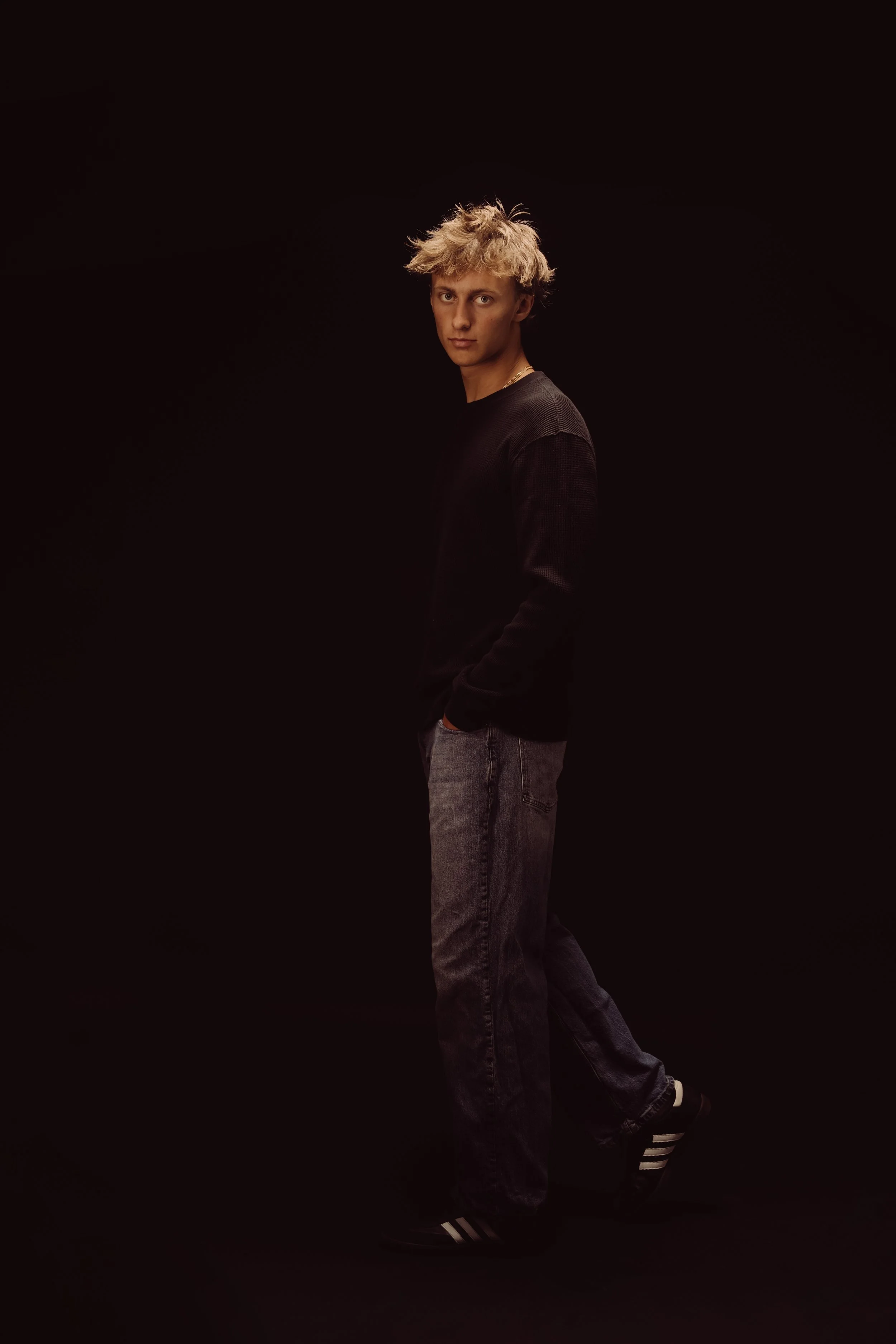 Young man standing against a black background, wearing a black long-sleeve shirt, gray jeans, and black Adidas sneakers with white stripes, with his hands in his pockets and looking at the camera.