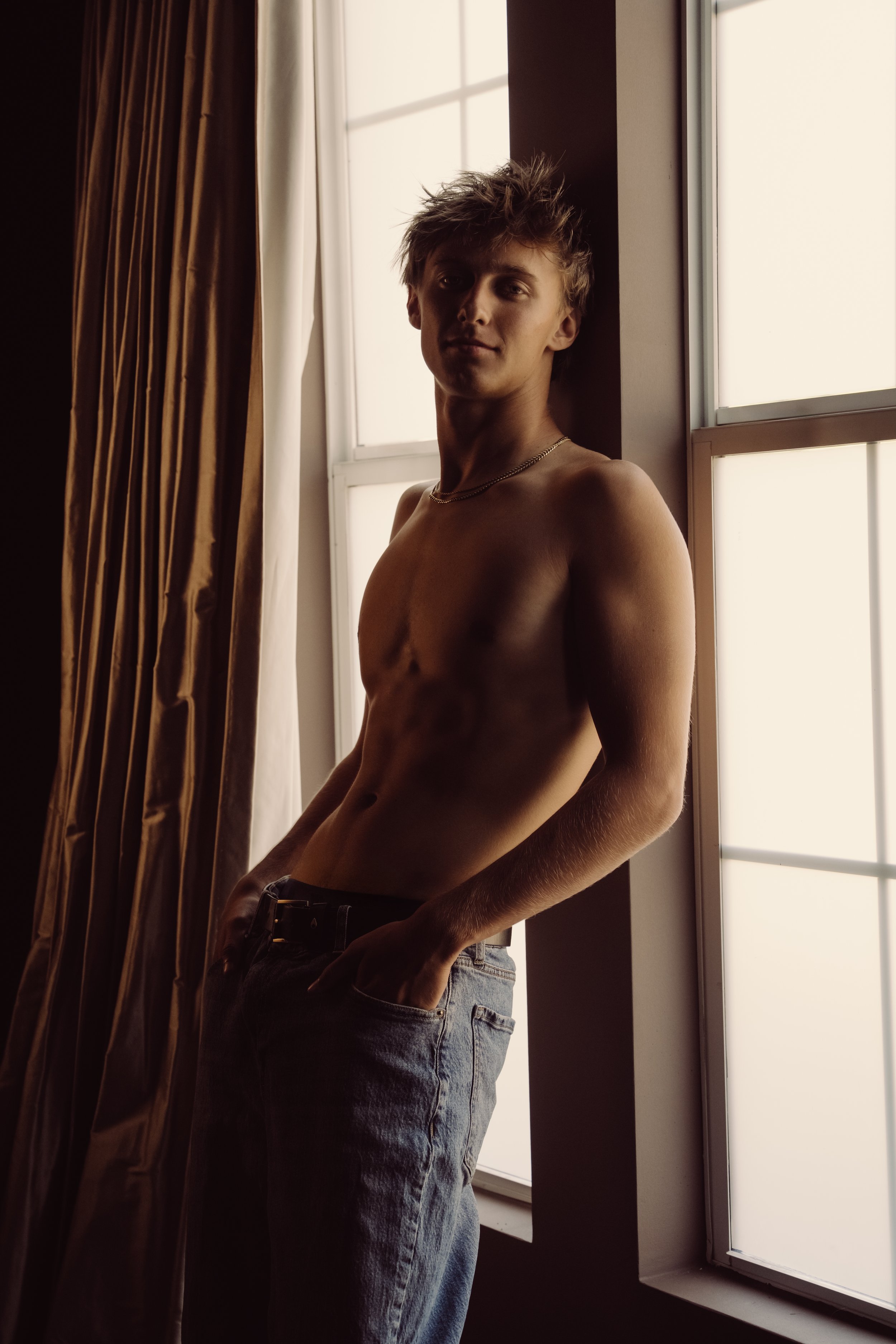 A young man with light skin and tousled hair standing shirtless near a window, wearing jeans, with a slight smile, in natural light.