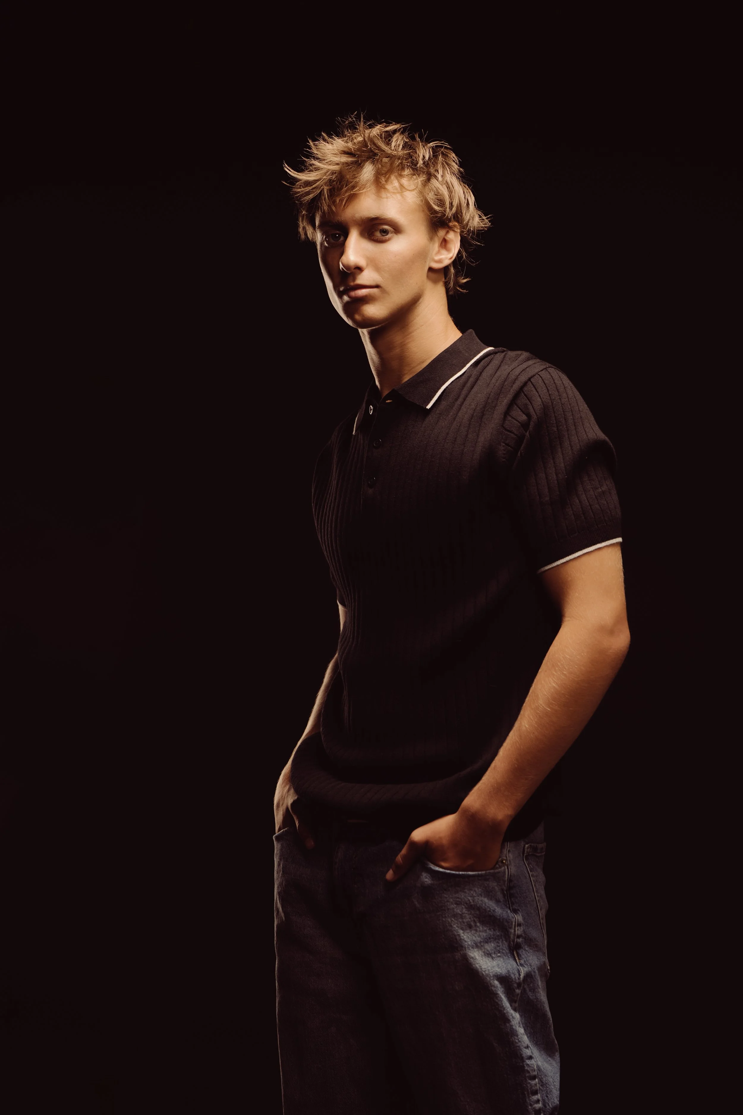 A young man with tousled blonde hair wearing a black polo shirt and jeans, standing against a dark background.