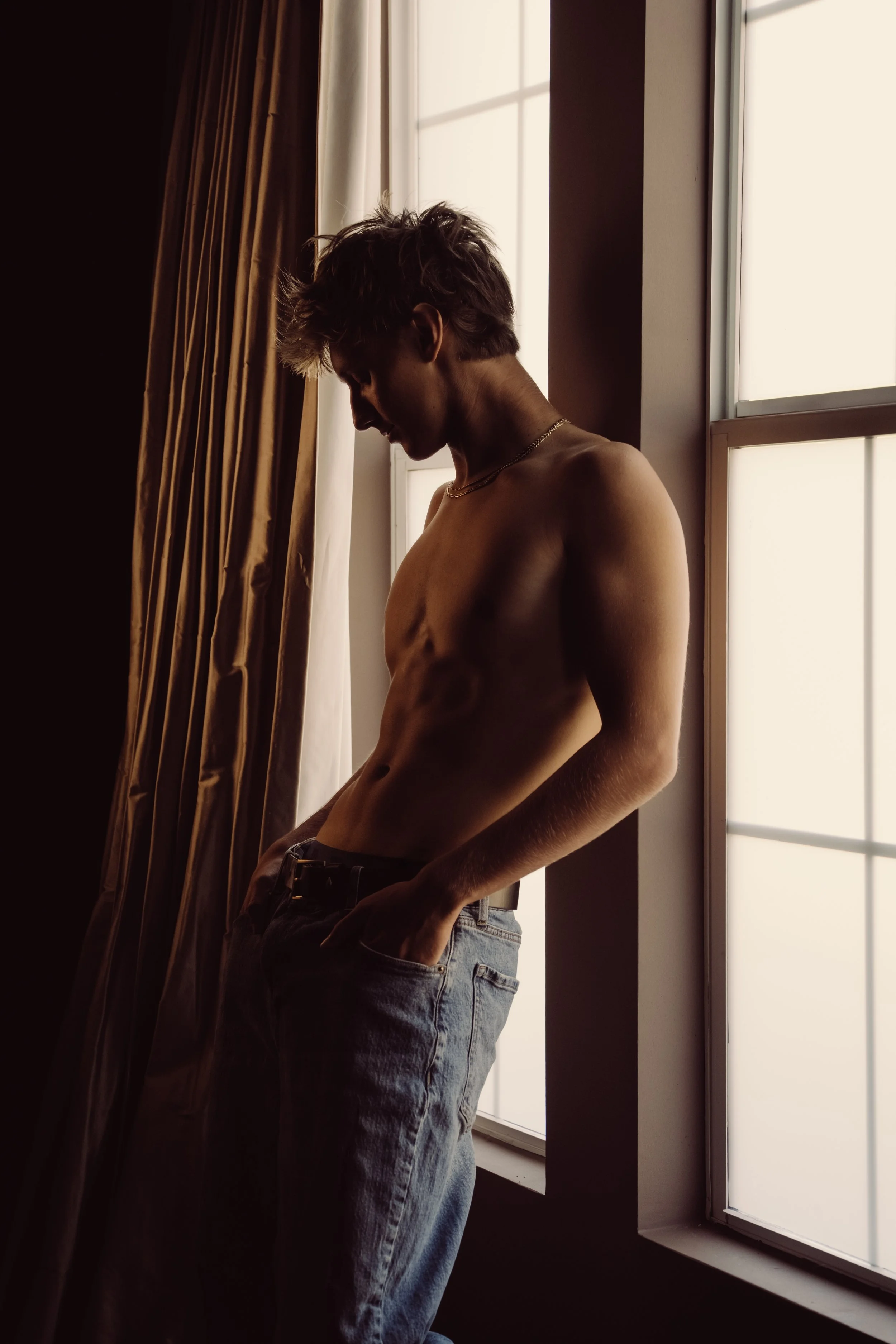 A shirtless man with disheveled hair standing by a window with light streaming in, wearing jeans with his hands in his pockets.