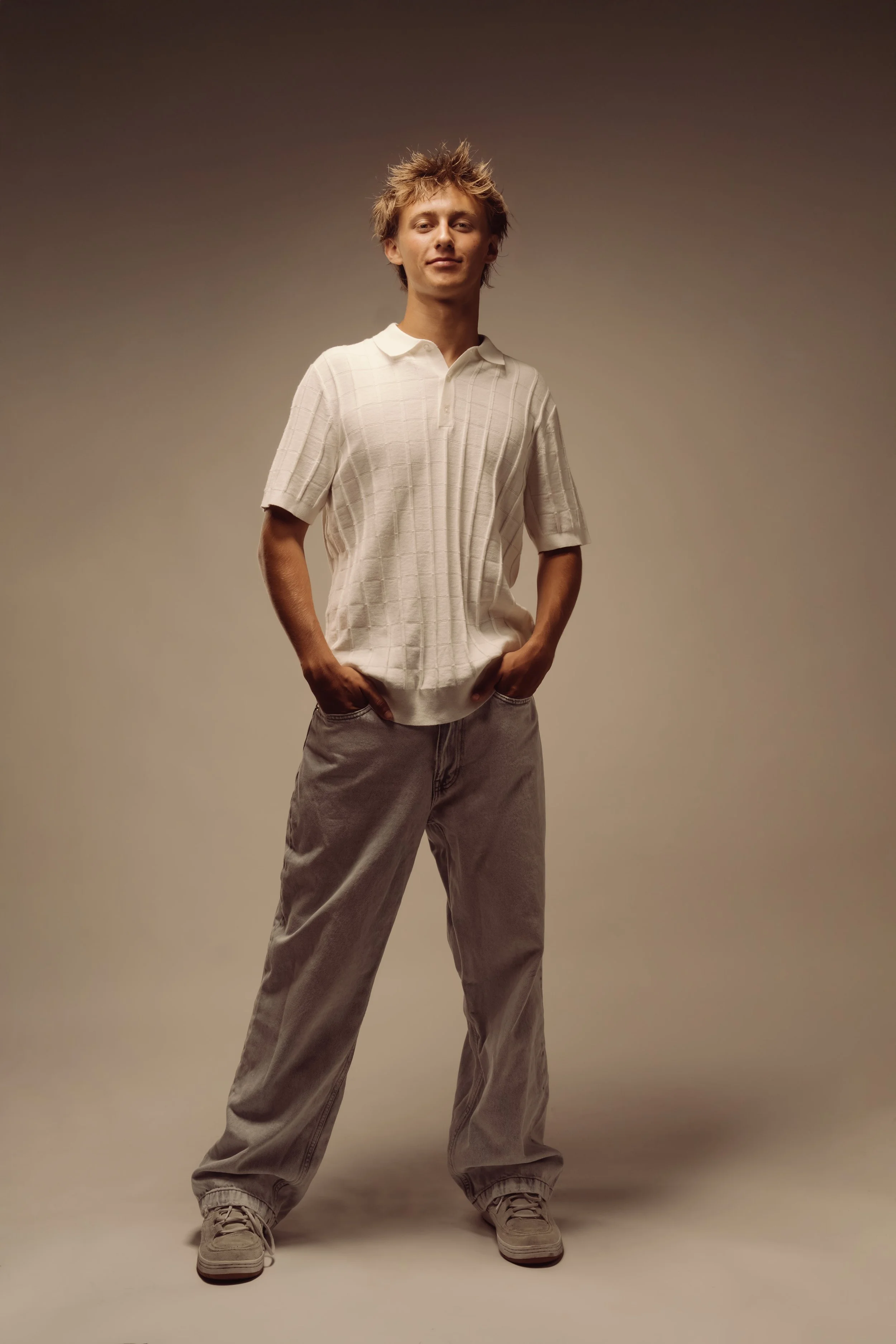 A young man with messy blonde hair stands confidently with hands in pockets, wearing a beige short-sleeved polo shirt and loose beige pants, against a neutral background.