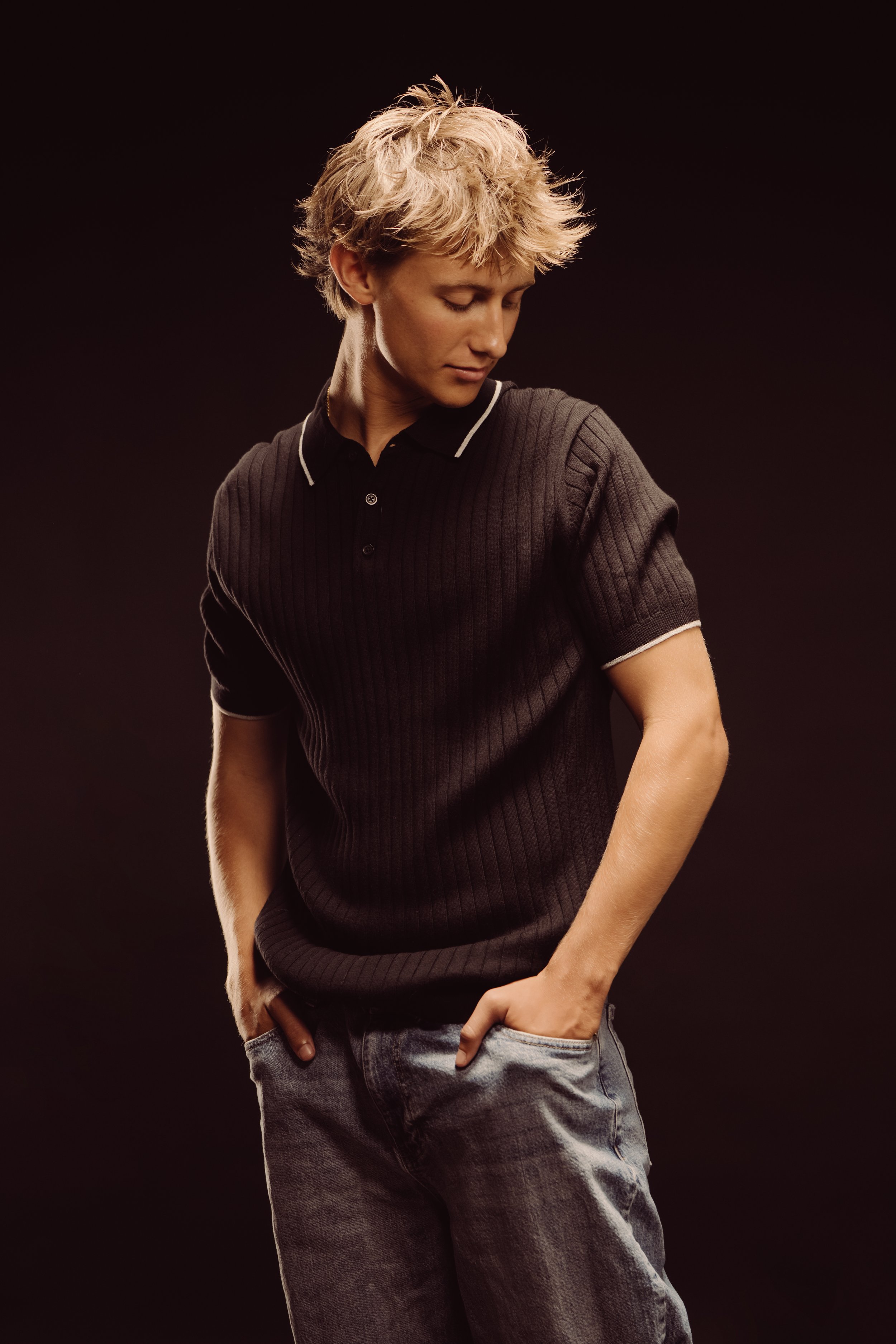 A young man with short, wavy blonde hair looking down, wearing a black polo shirt and faded jeans against a dark background.