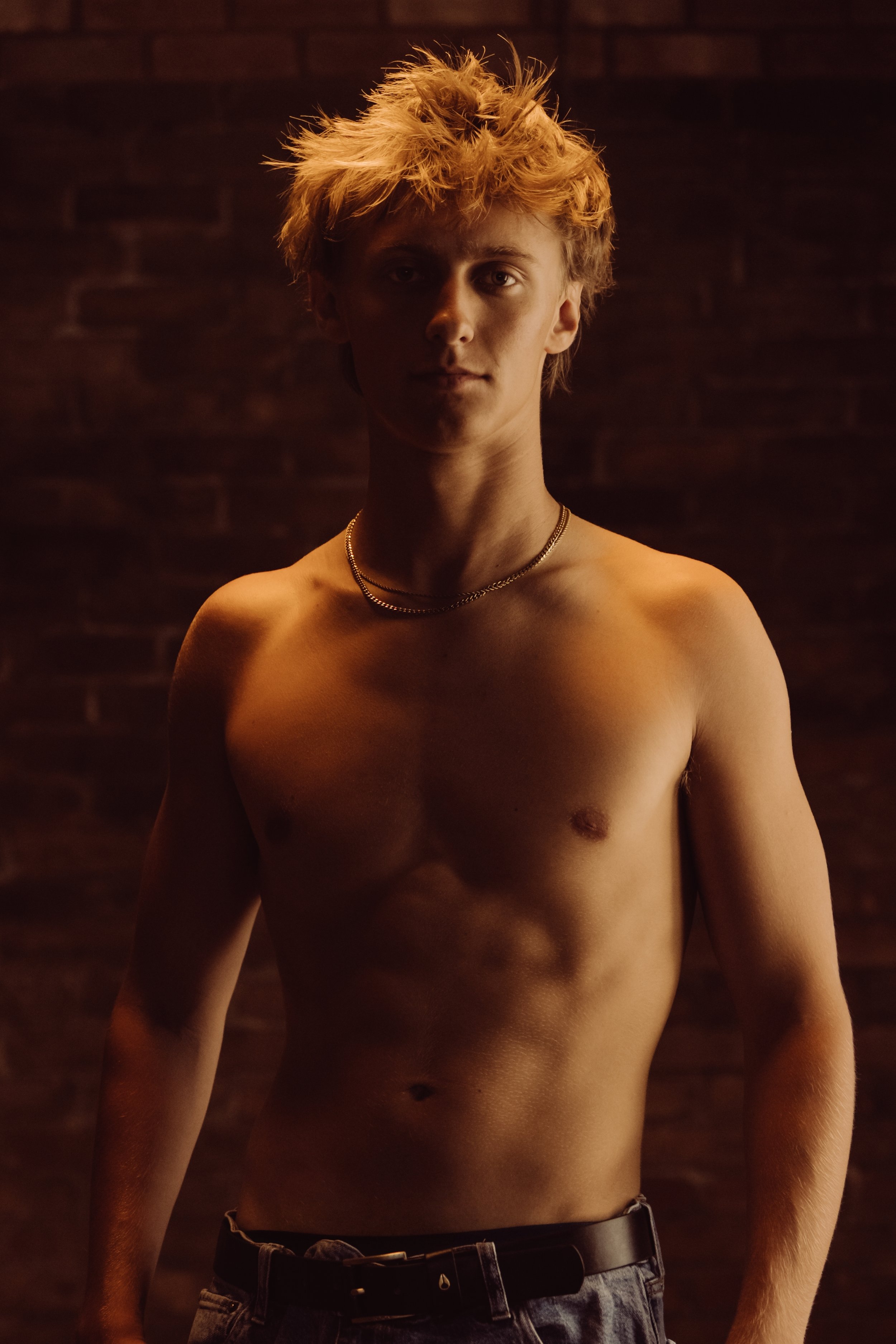 A shirtless young man with messy blonde hair is standing in front of a brick wall, with low lighting highlighting his face and upper body.