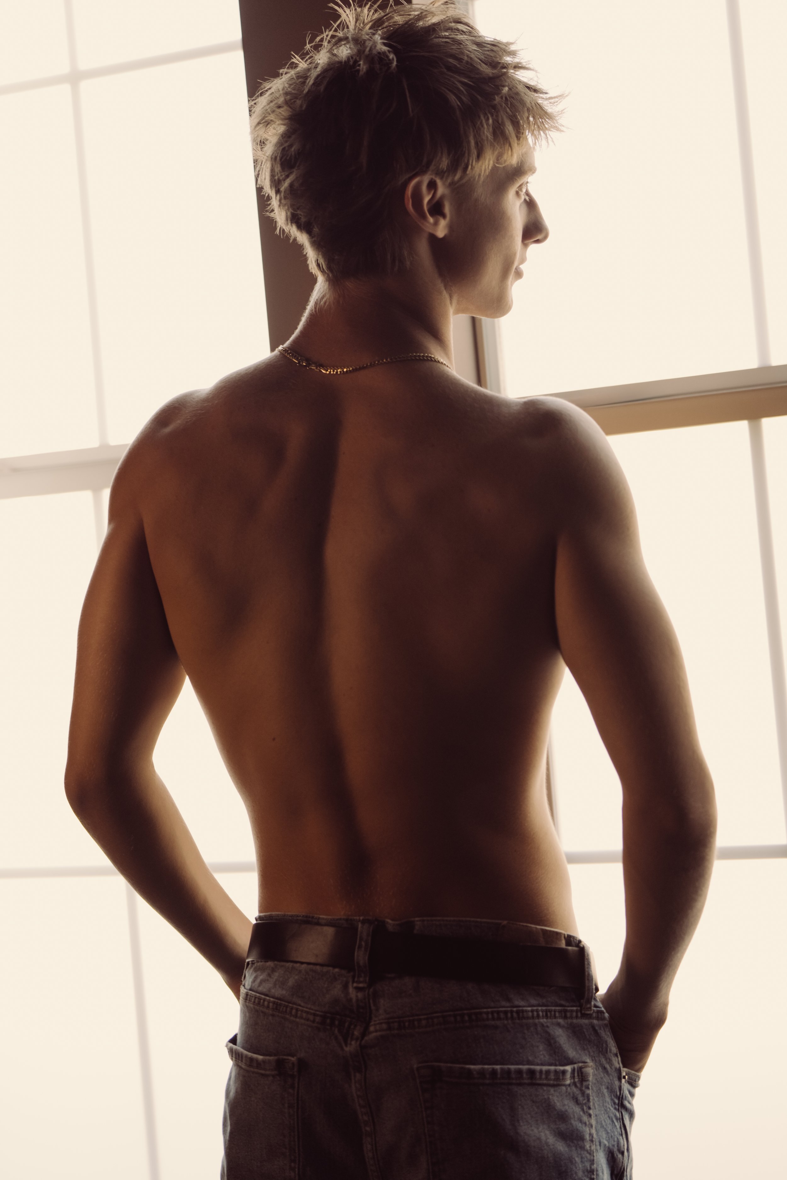 Back view of a young man with a muscular build, blonde hair, wearing a gold chain, shirtless, standing near a window with daylight filtering through.