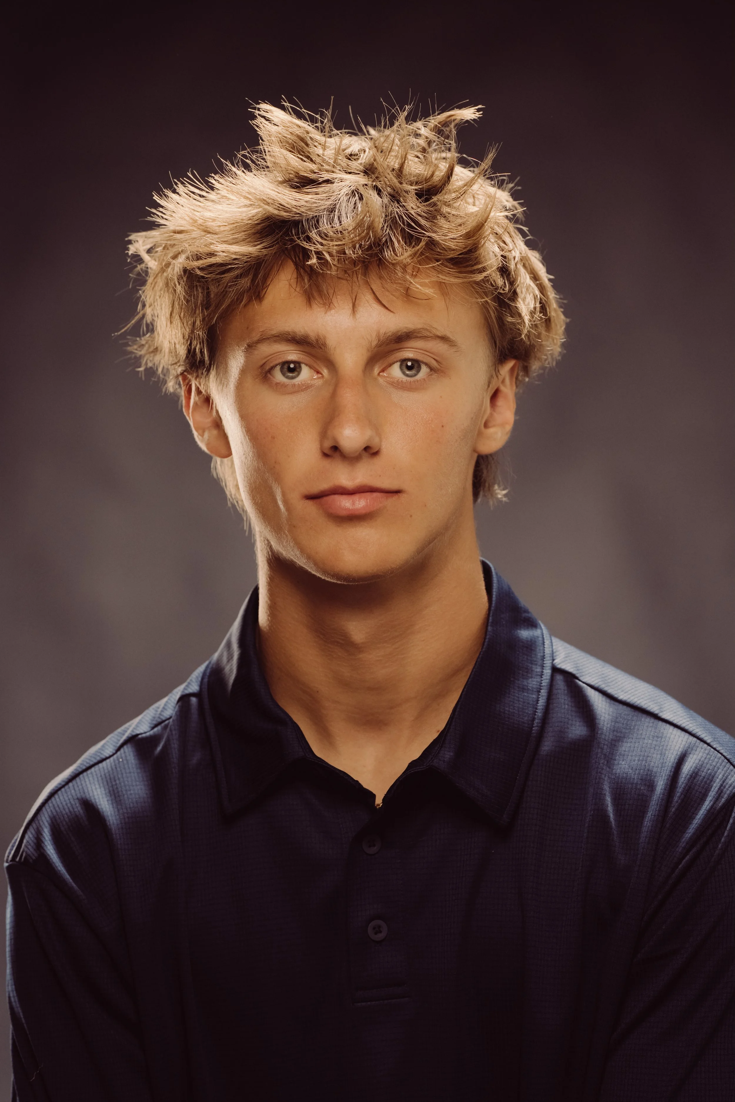 Portrait of a young man with voluminous, tousled blond hair, blue eyes, and light skin, wearing a dark collared shirt against a gray background.