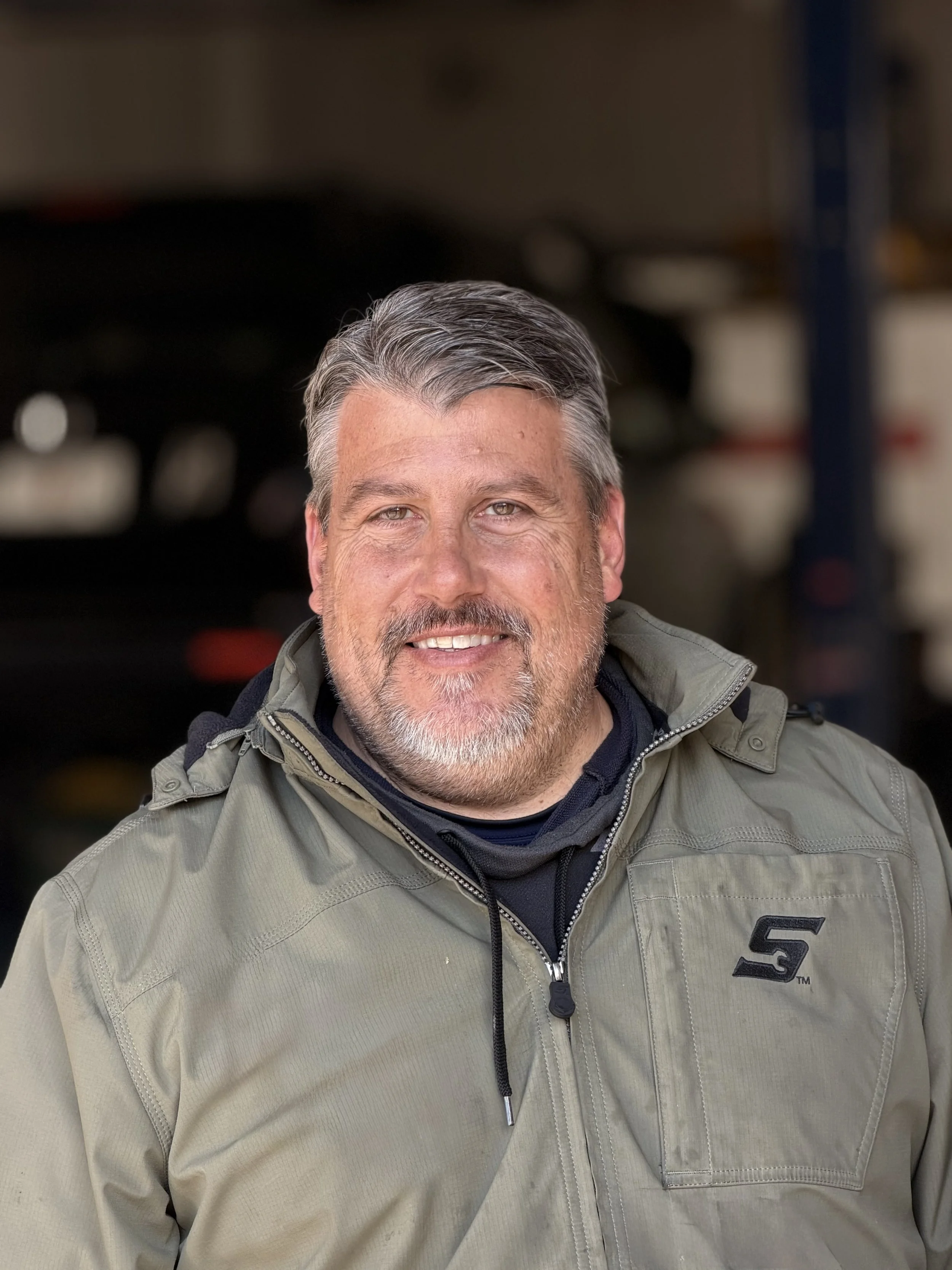 Owner Ken smiling at the camera, wearing a beige jacket with a black logo on the chest, in a garage or workshop environment.