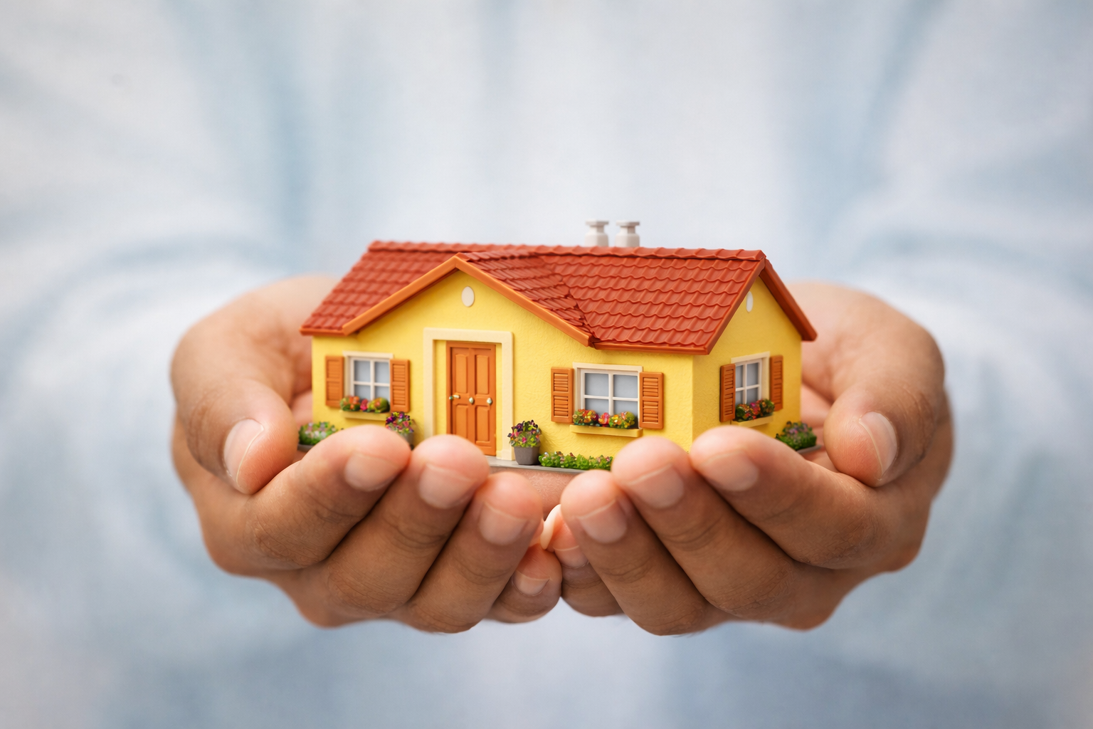 adult-residential-facility | A person holding a miniature yellow house with red roof shingles, brown window shutters, and flower boxes with colorful flowers.