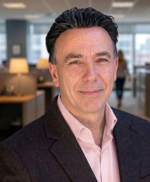 A middle-aged man with dark hair, wearing a pink shirt and dark blazer, smiling in an office environment.