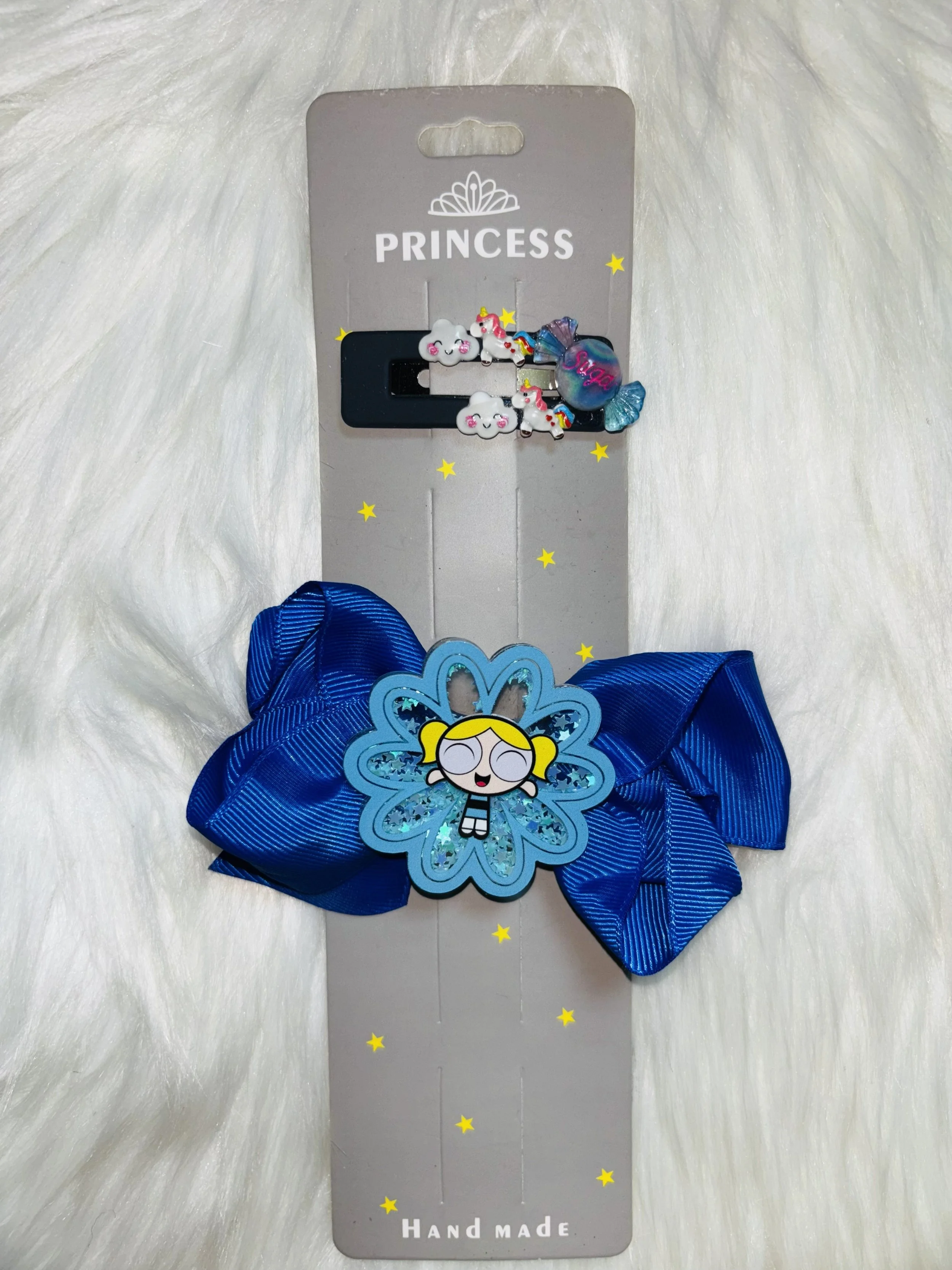 Hair clip with a blue bow, a character with a blonde ponytail with a crown, and a snack-shaped charm, all attached to a gray card labeled 'Princess' and 'Hand Made,' with small yellow stars on a white furry background.
