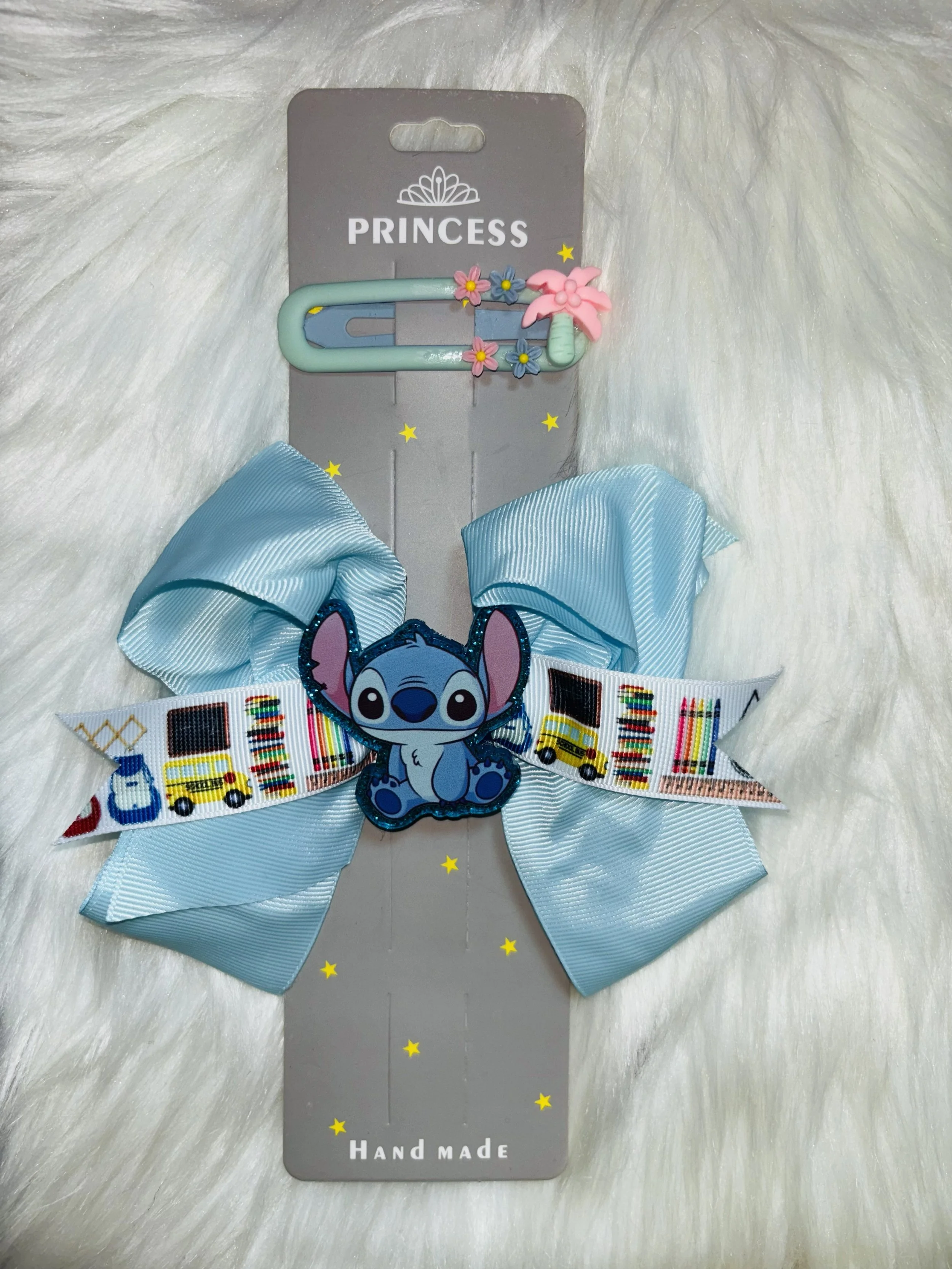 A decorative hair clip with blue and pink elements, including an image of Stitch from Disney's Lilo & Stitch, attached to a gray display card with the words 'PRINCESS' and 'Hand Made' and small yellow star decals on a white furry background.