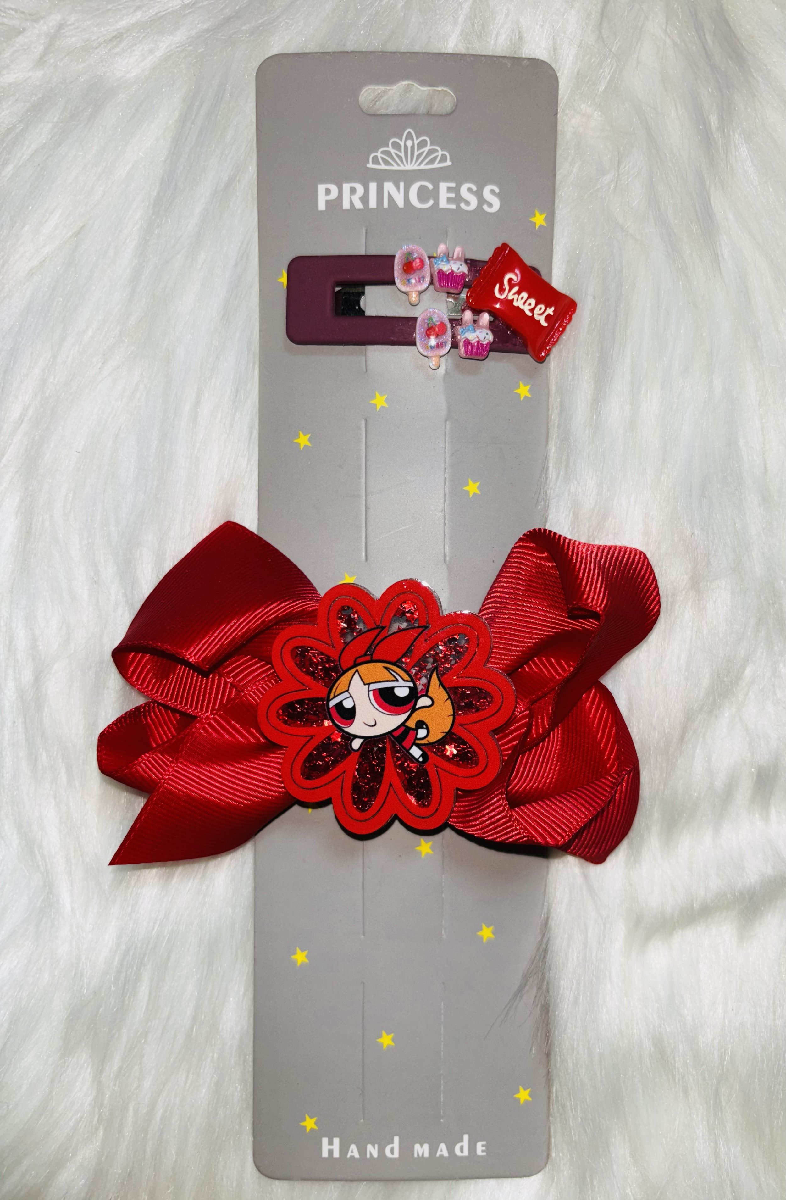 A handmade hair bow featuring a red ribbon, a decorative flower with a cartoon character, and small pink and purple cupcake charms attached to a cardboard backing labeled 'PRINCESS' and 'Hand Made'.