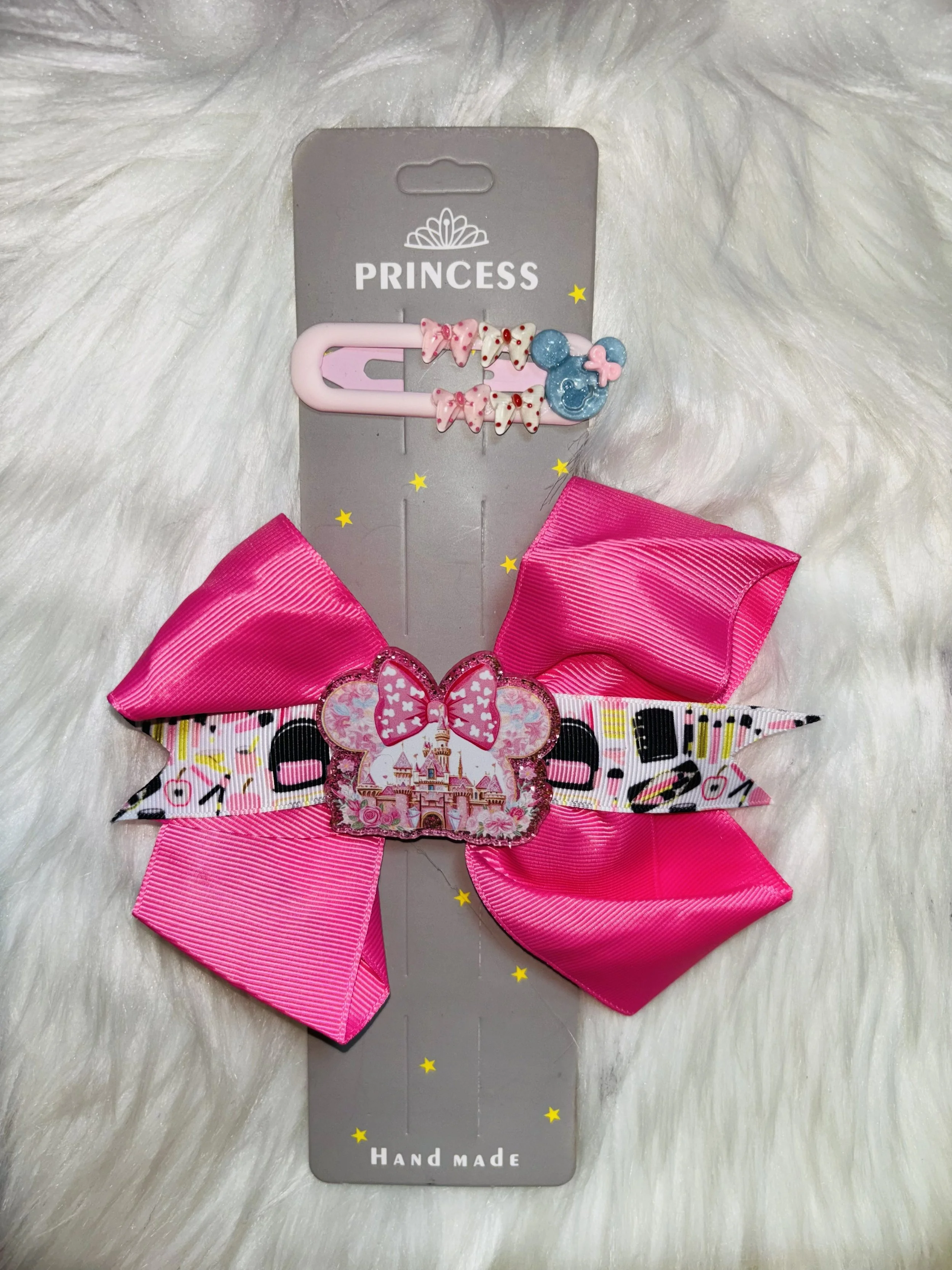 A pink safety pin decorated with small pink and white bows and a blue Mickey Mouse head with a pink bow, attached to a gray card with "PRINCESS" written on it, and a large pink bow at the bottom, all on a white fluffy background.