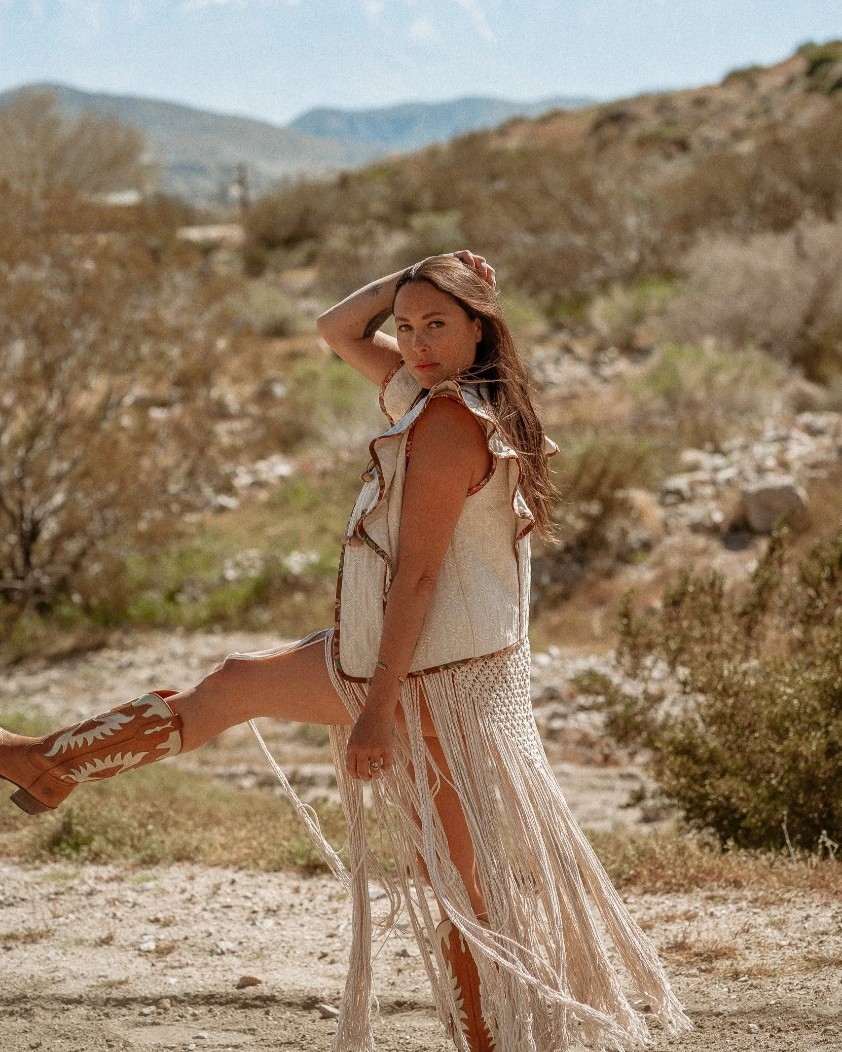 A young woman standing outdoors in a desert landscape with mountains in the background, wearing a cream-colored fringed skirt, matching top, and cowboy boots.