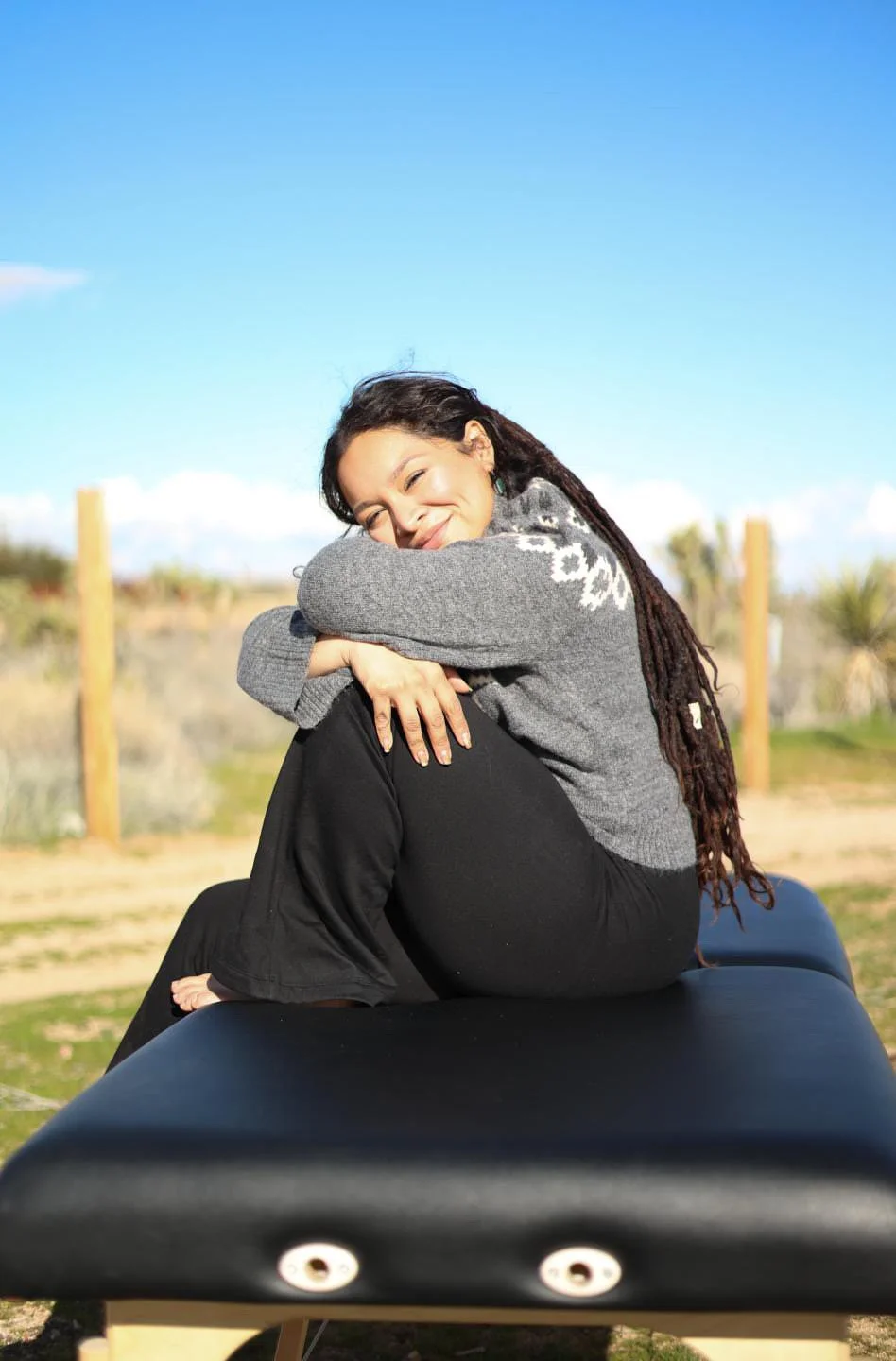 Young woman with dreadlocks sitting outdoors on a black bench, smiling and hugging her knees, wearing a gray sweater with white patterns, with a blue sky and desert plants in the background.